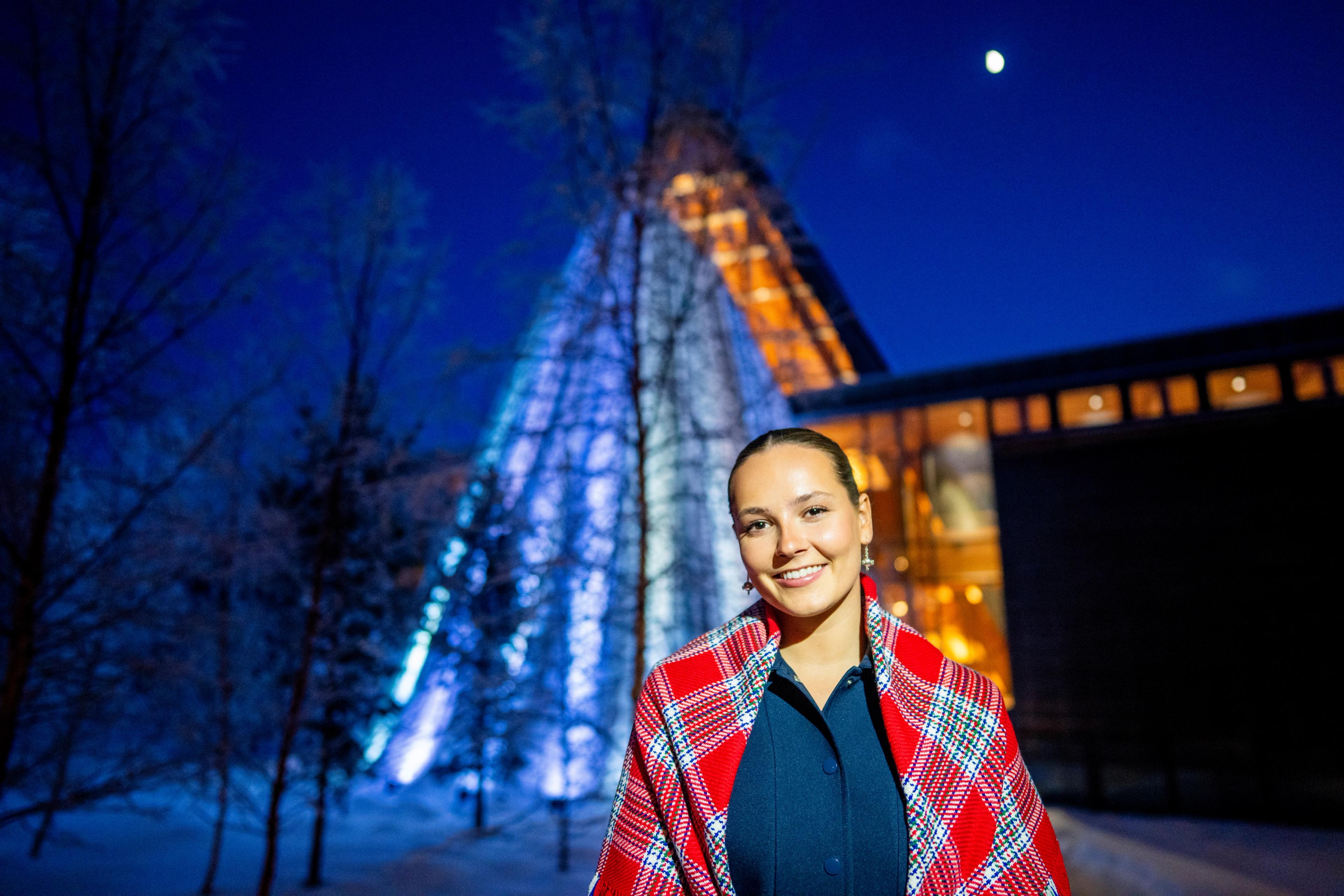 Tett på Prinsesse Ingrid Alexandra i mørke ute, ikledd samisk sjal og med en lyssatt sametingsbygning bak.