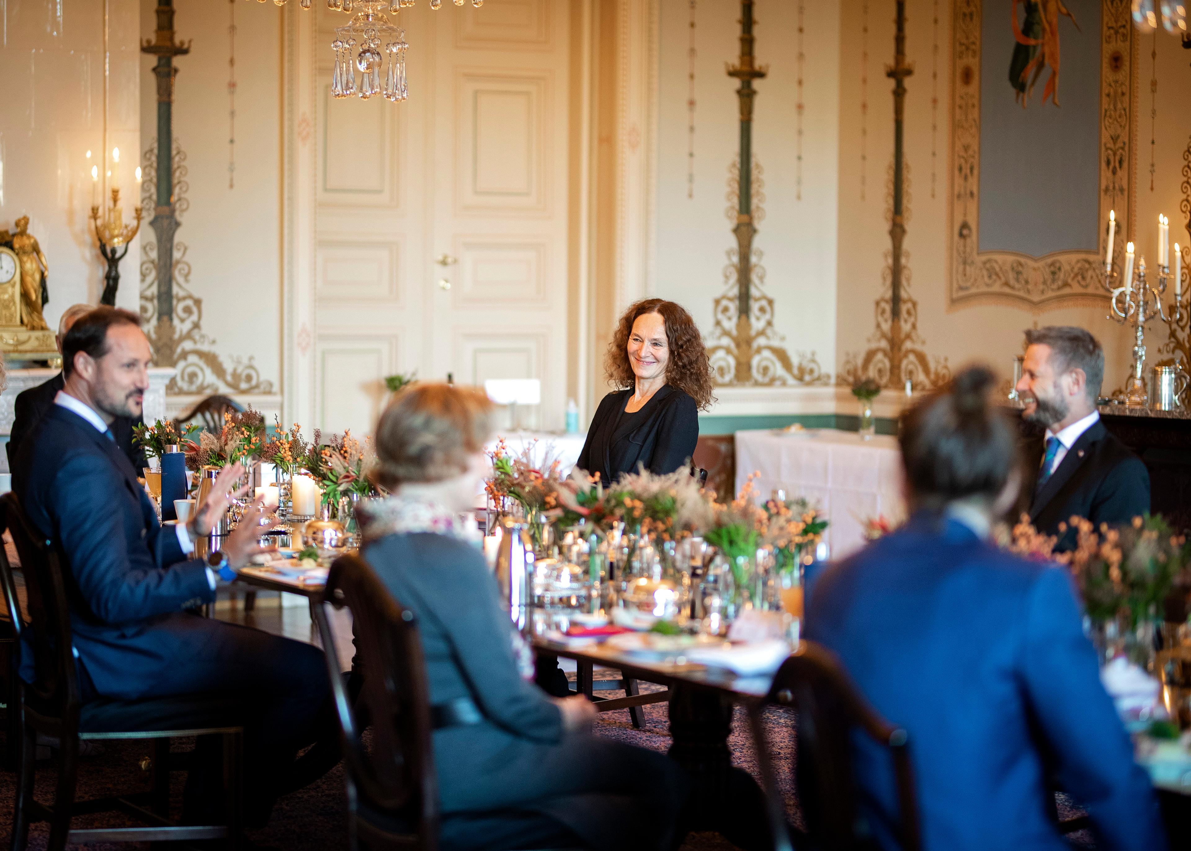 Helseminister Bent Høie og Camilla Stoltenberg fra Folkehelseinsituttet lytter til Kronprinsen. Sitter rundt et dekket bord i Daglige spisesal, med "koronaavstand" mellom plassene.