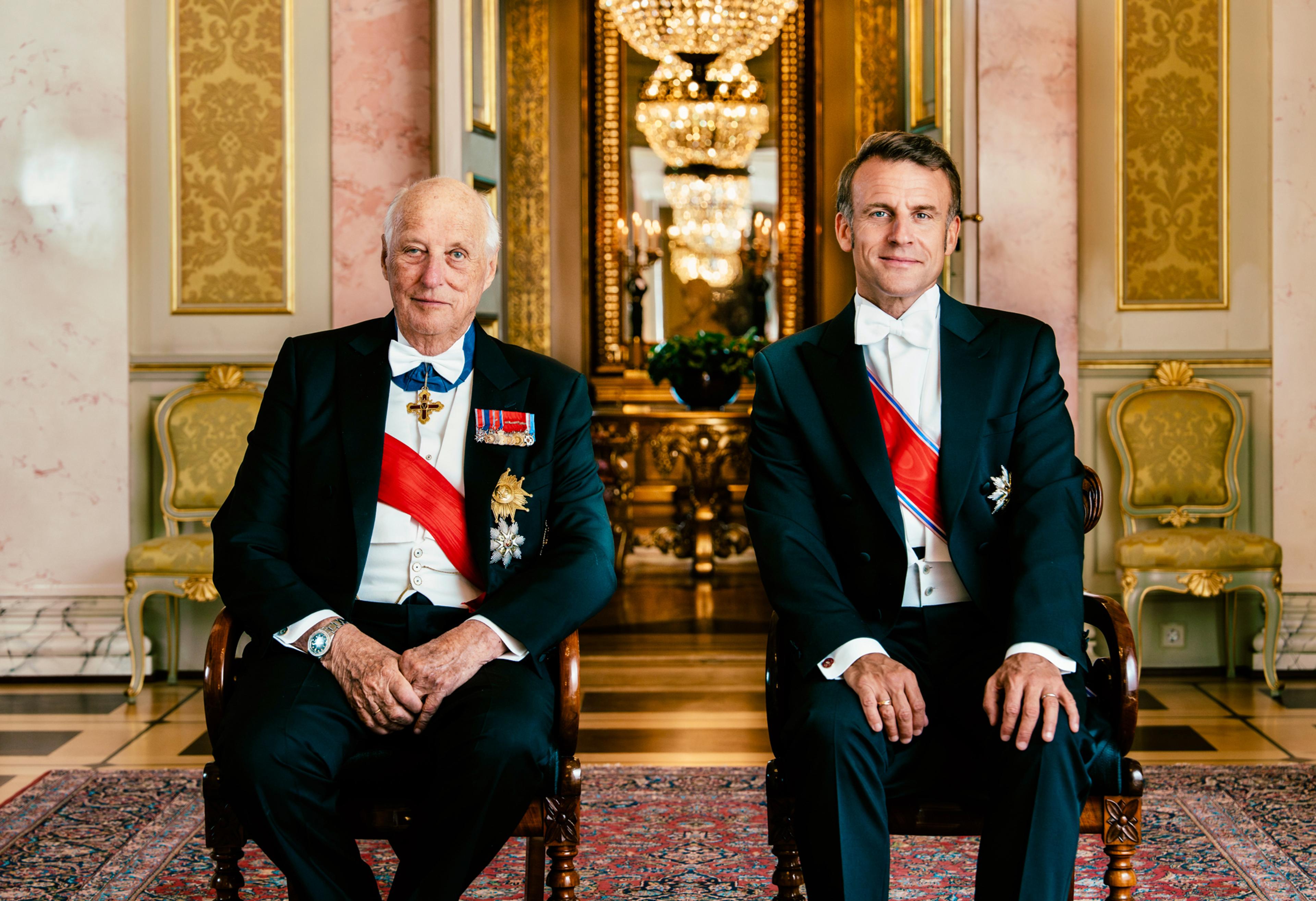 Kong Harald og Frankrikes president Emmanuel Macron sittende ved siden av hverandre. Begge ser direkte inn i kameraet.