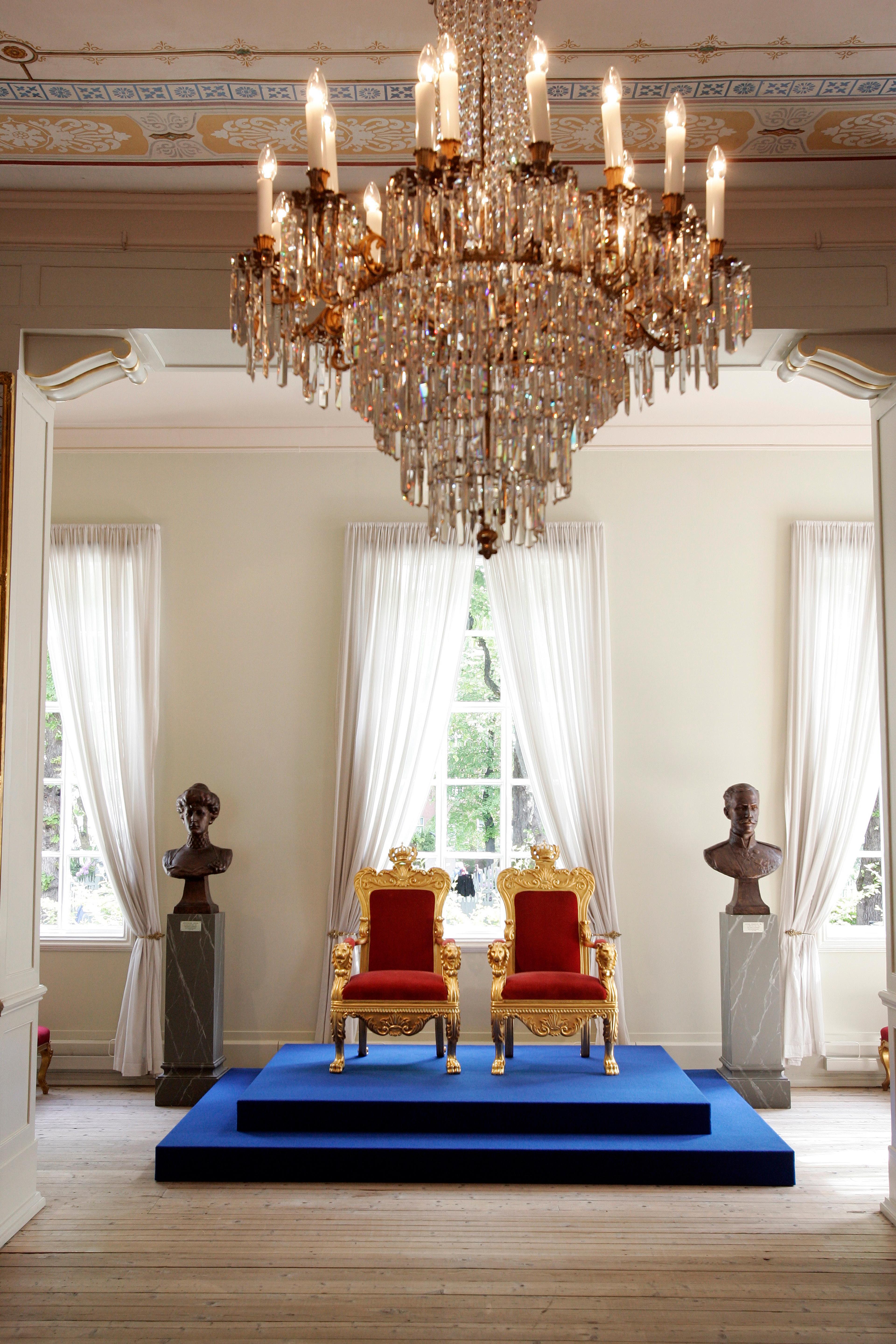 To gullforgylte stoler med seter i rød fløyel og kroner på toppen av ryggen står på et blått podium i et vakkert rom.