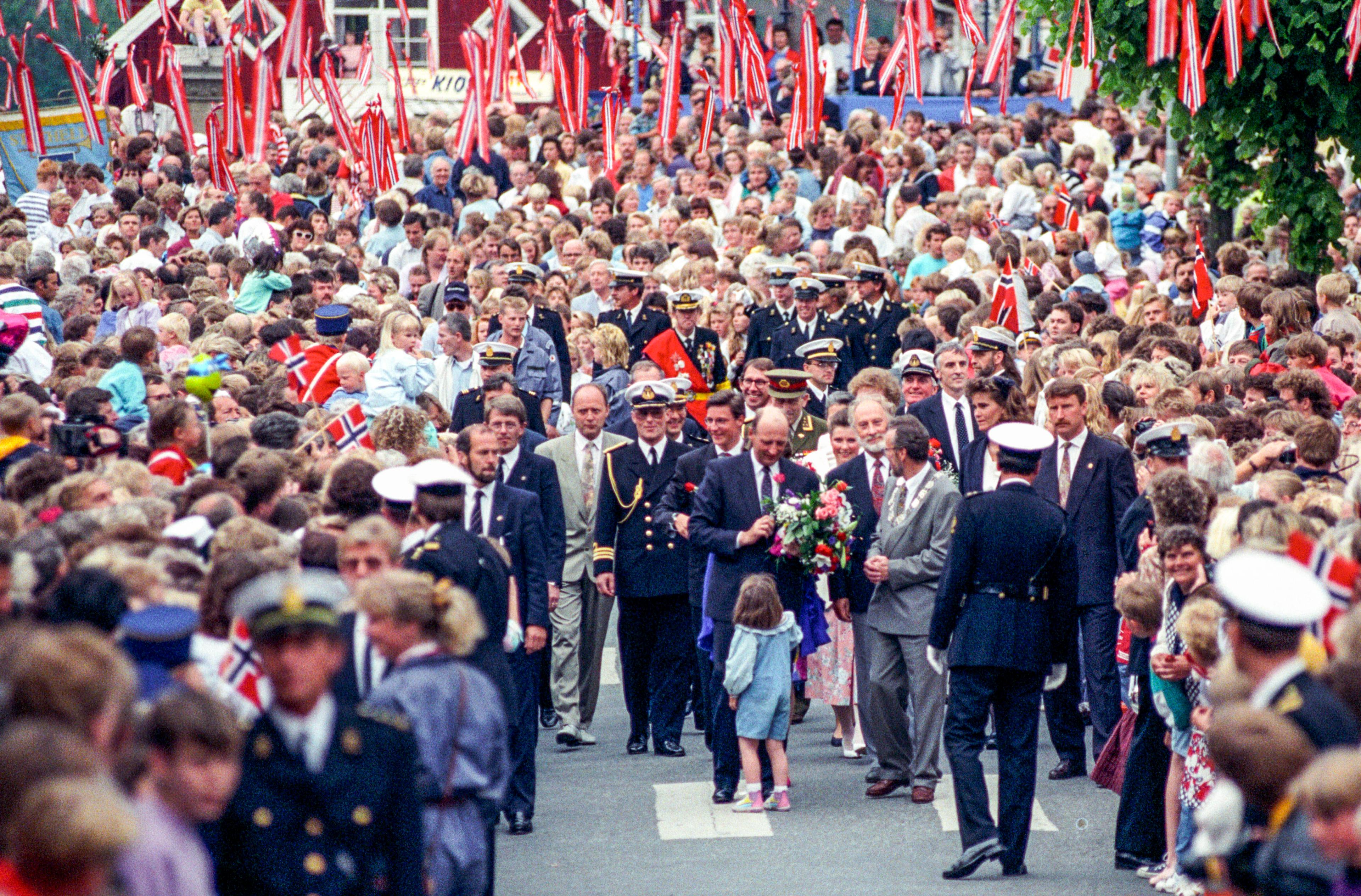 Kong Harald og blomsterpiken på gaten, omkranset av en stor menneskemengde. Det er pyntet med norske flagg.