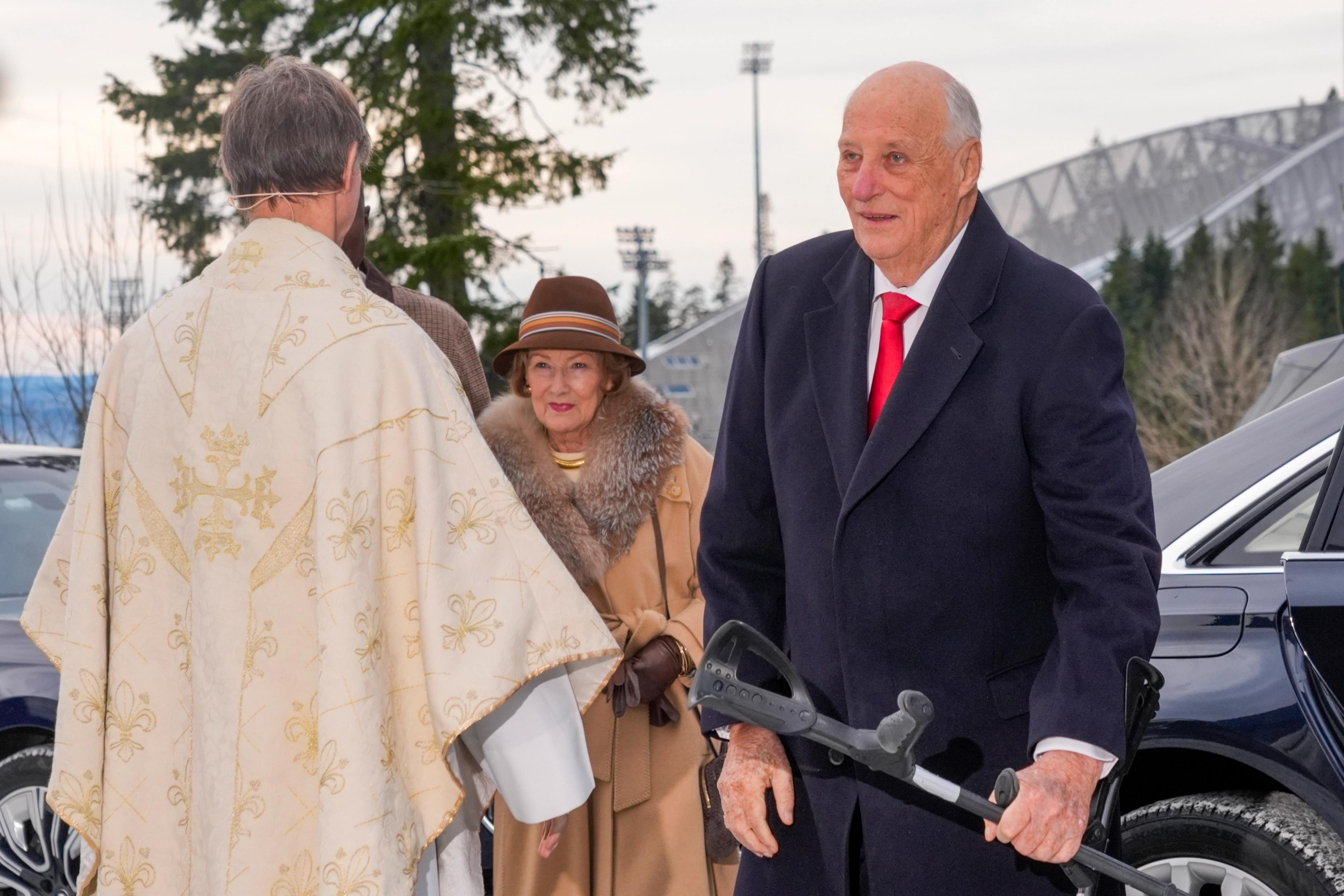 ong Harald går soknepresten i møte. Dronning Sonja følgjer like bak. Vi ser delar av Holmenkollbakken i bakgrunnen.