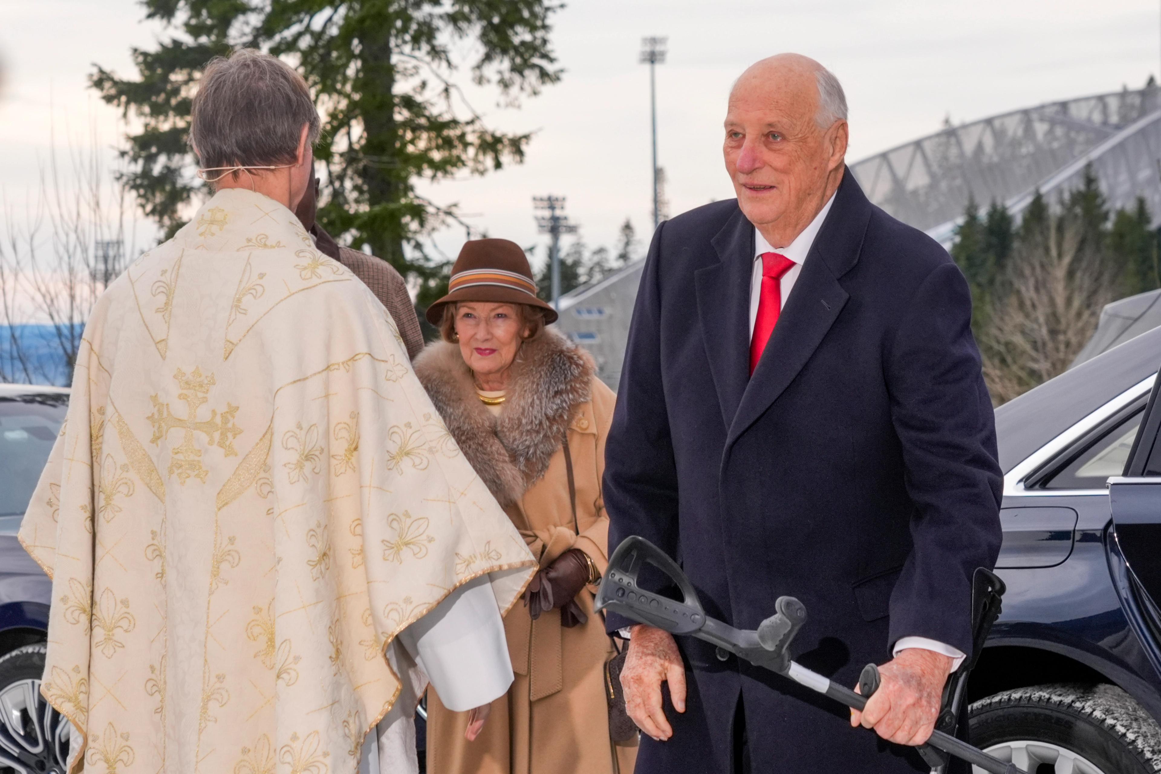 ong Harald går soknepresten i møte. Dronning Sonja følgjer like bak. Vi ser delar av Holmenkollbakken i bakgrunnen.