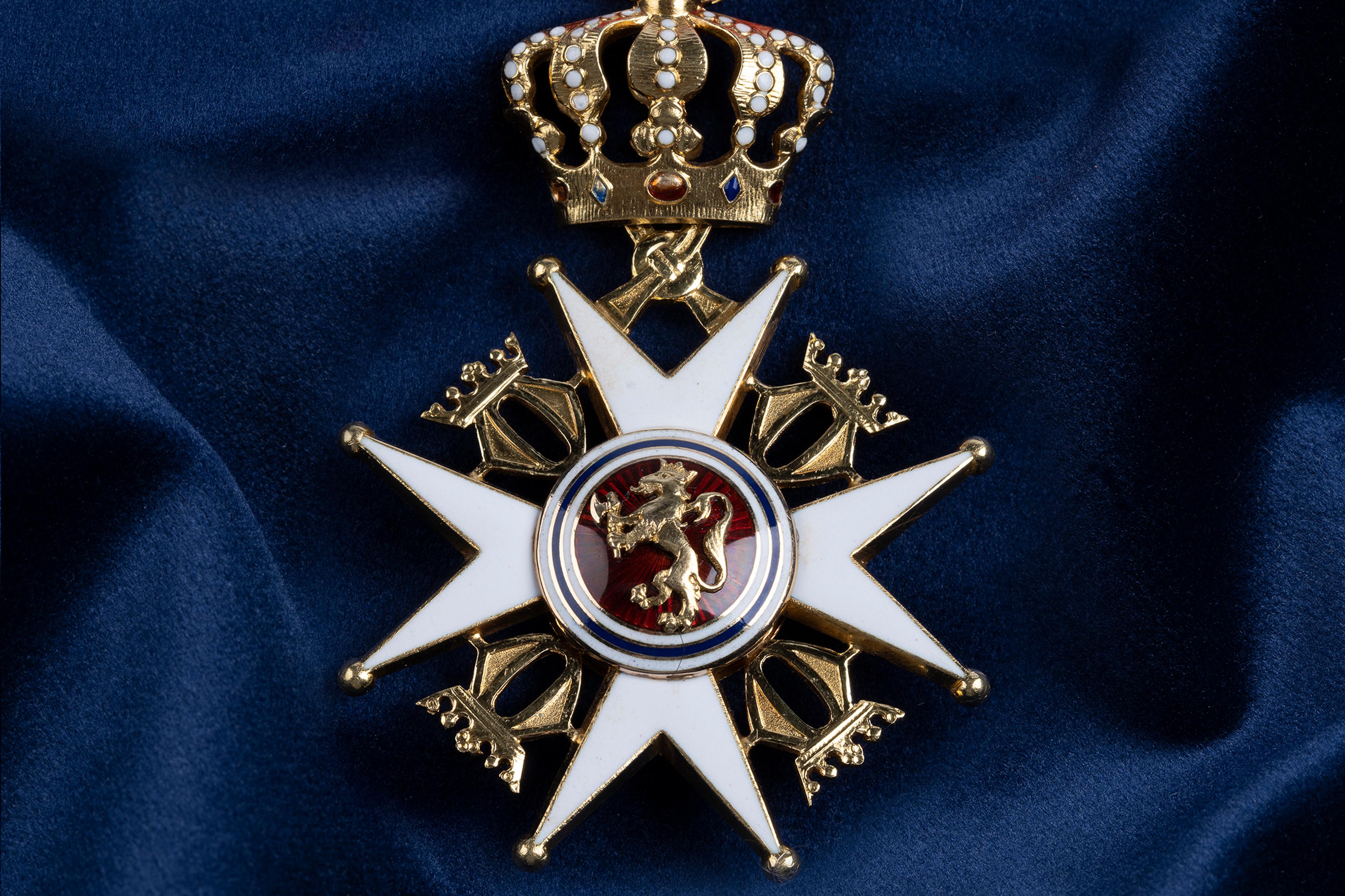 Nærbilde av ordensmerket festet i rødt bånd med blå og hvite striper på en bakgrunn av blått klede.