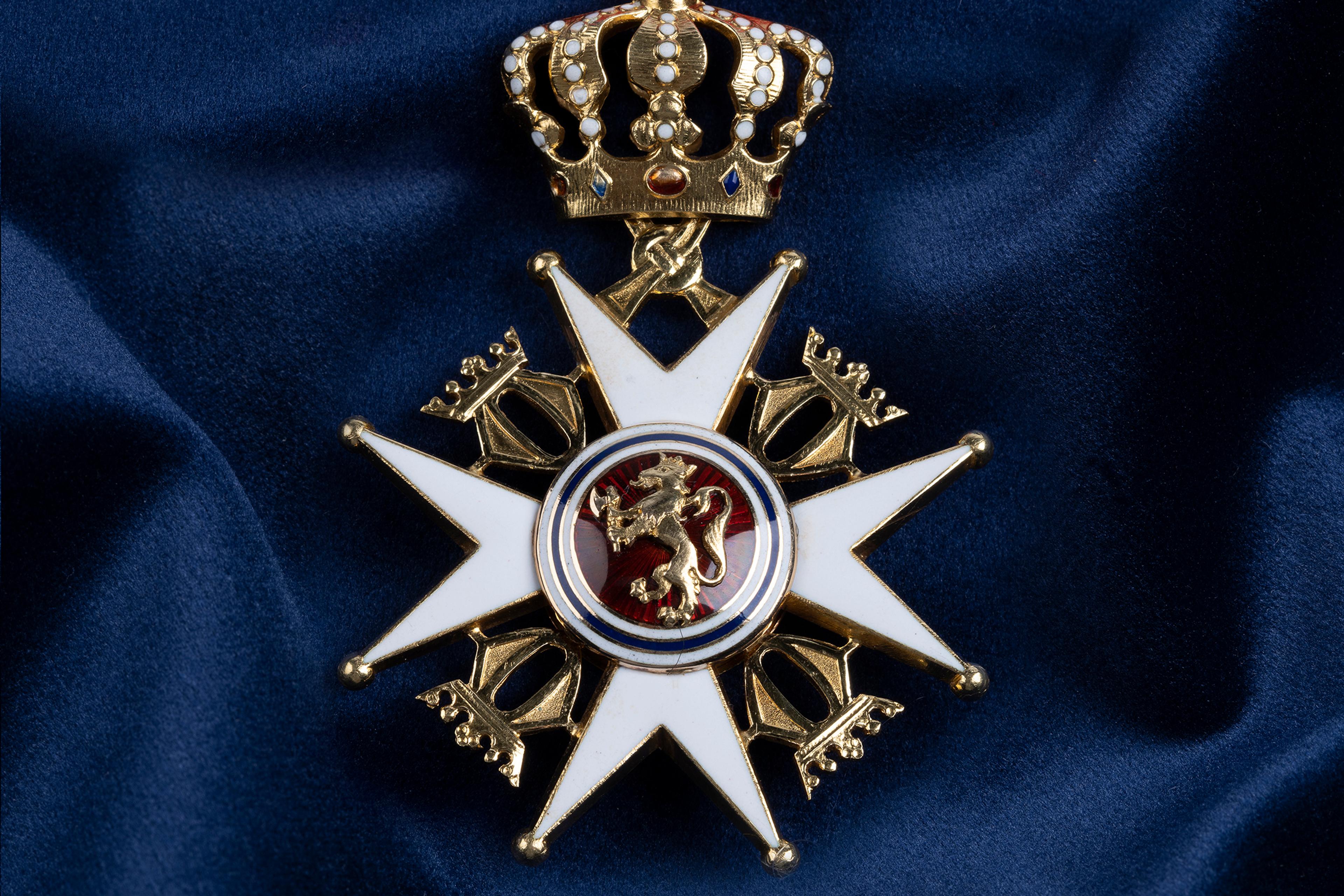 Nærbilde av ordensmerket festet i rødt bånd med blå og hvite striper på en bakgrunn av blått klede.