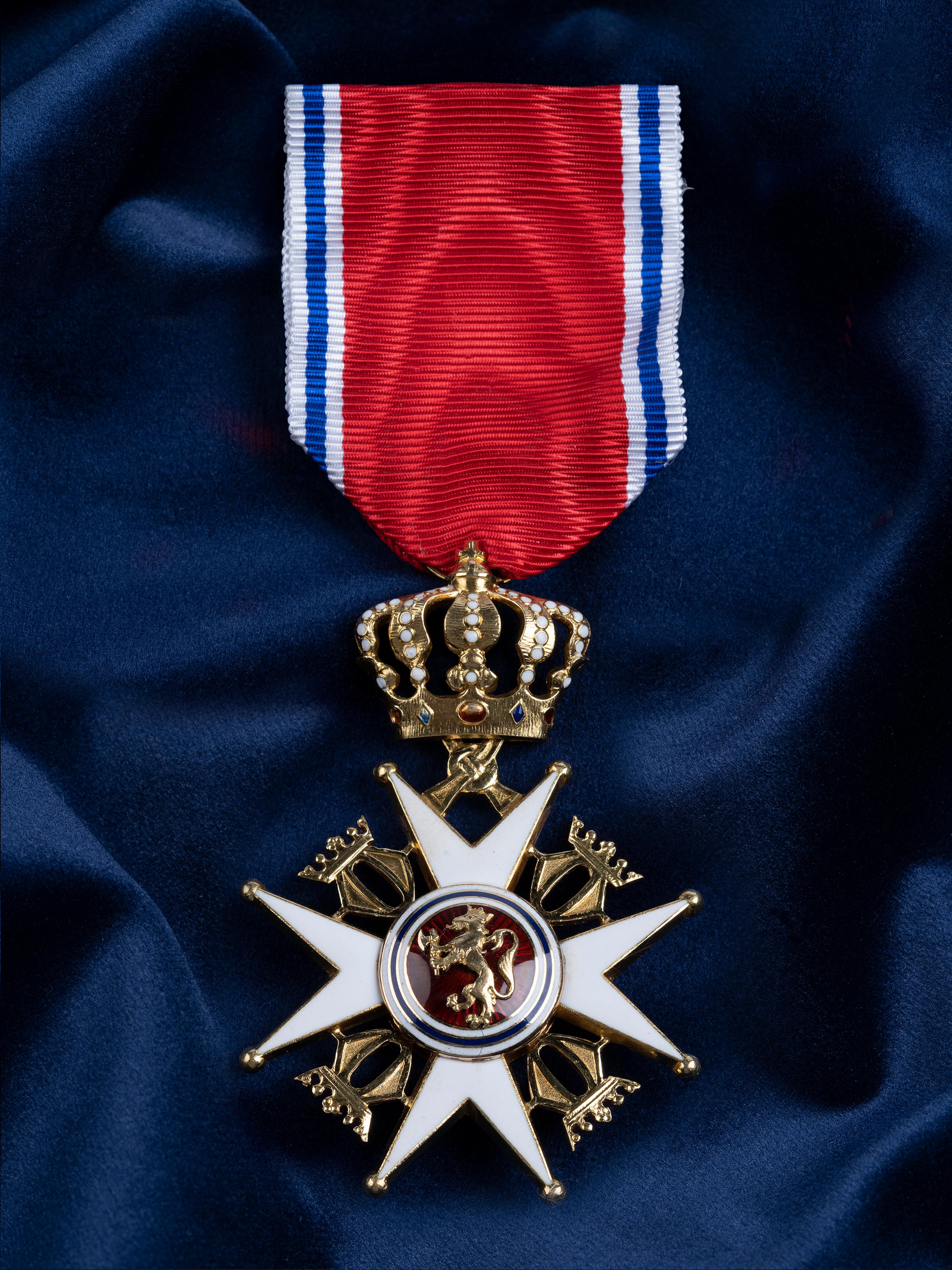 Nærbilde av ordensmerket festet i rødt bånd med blå og hvite striper på en bakgrunn av blått klede.