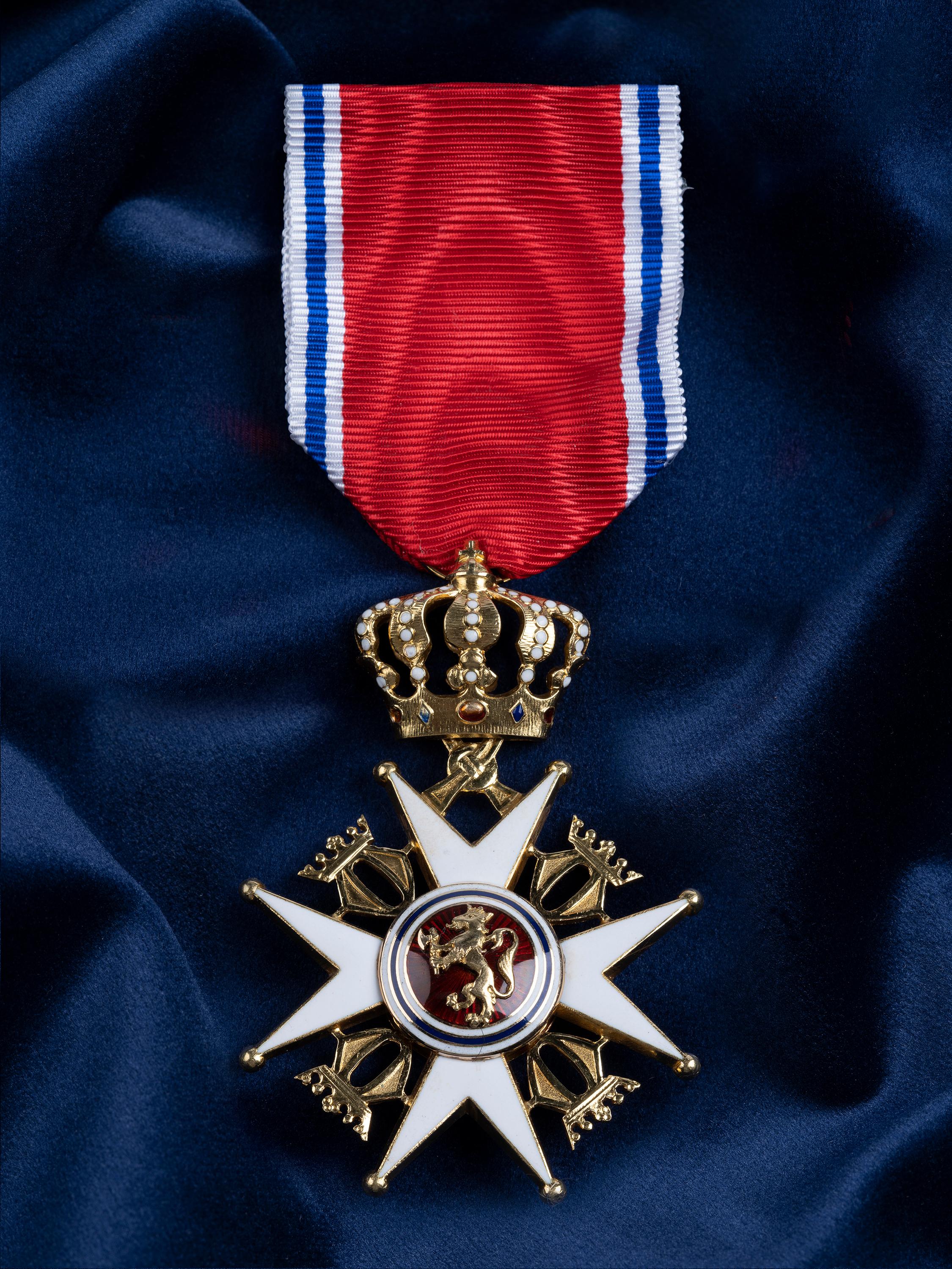 Nærbilde av ordensmerket festet i rødt bånd med blå og hvite striper på en bakgrunn av blått klede.