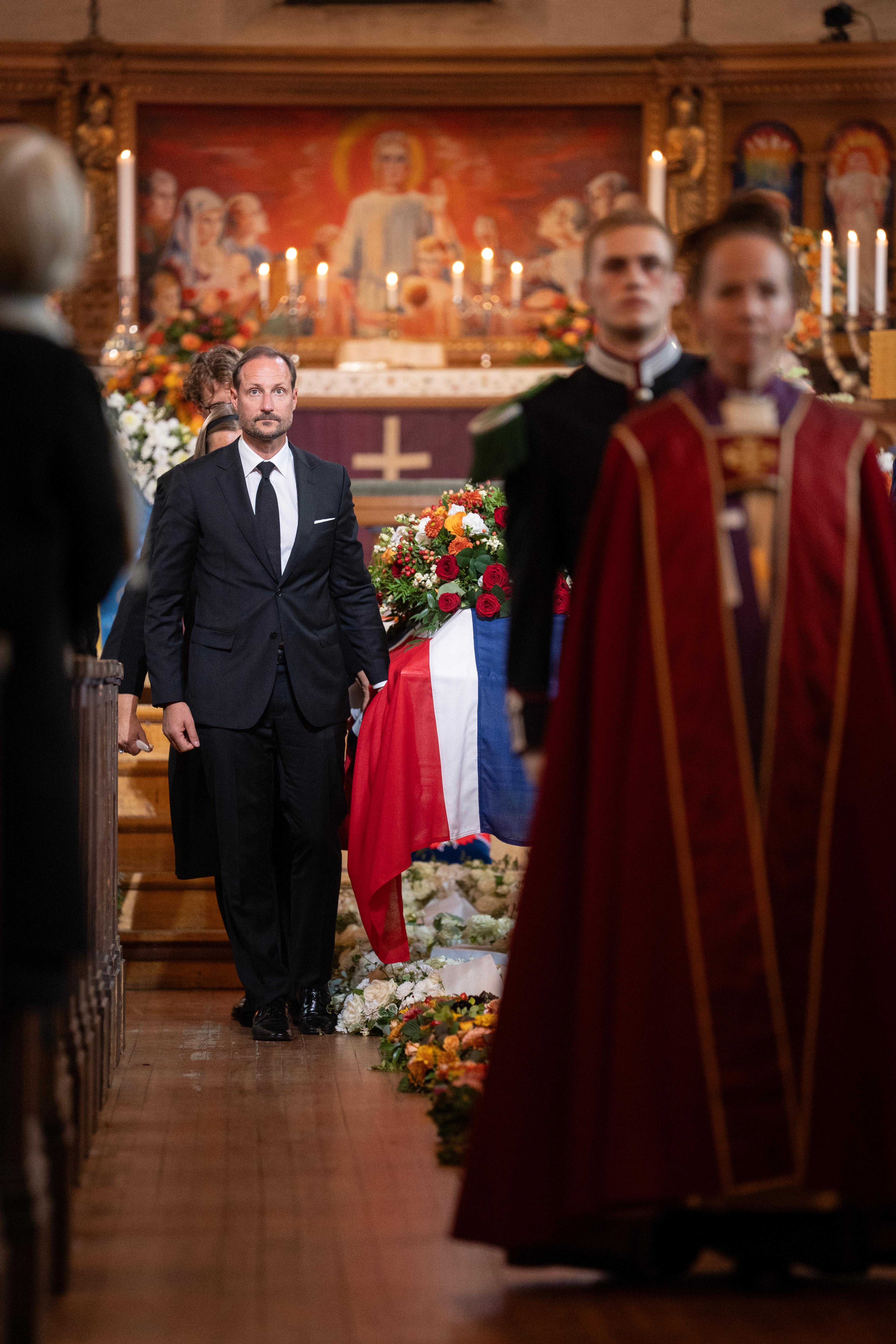Båren dekket med det norske flagget og en stor krans med fargerike blomstser bæres ned midtgangen i Ris kirke.