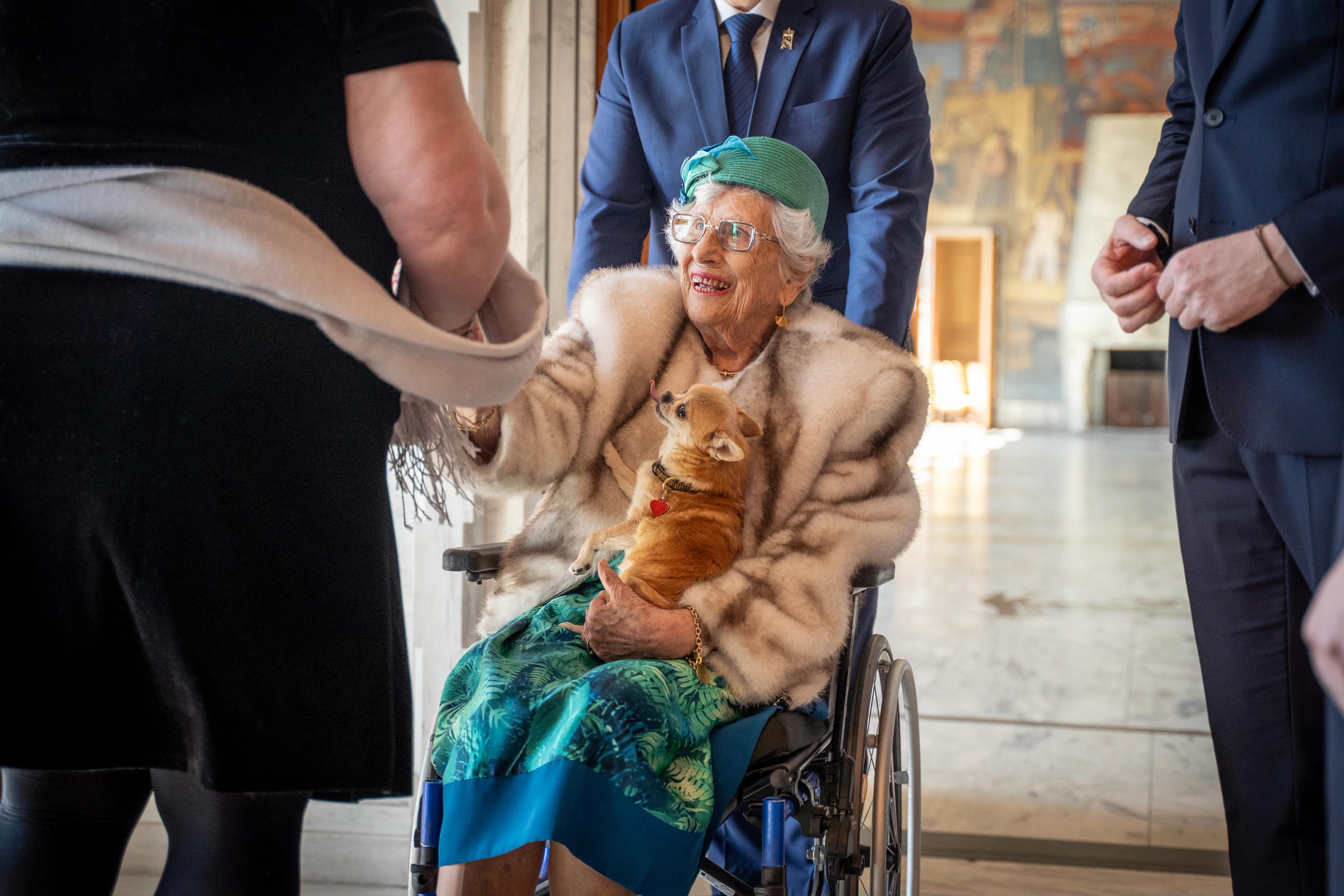 Prinsesse Astrid sitter i en rullestol med hunden Wilma på fanget. Hun rekker ut en hånd til styreleder Kathrin Dahlstrøm, som står med ryggen til.