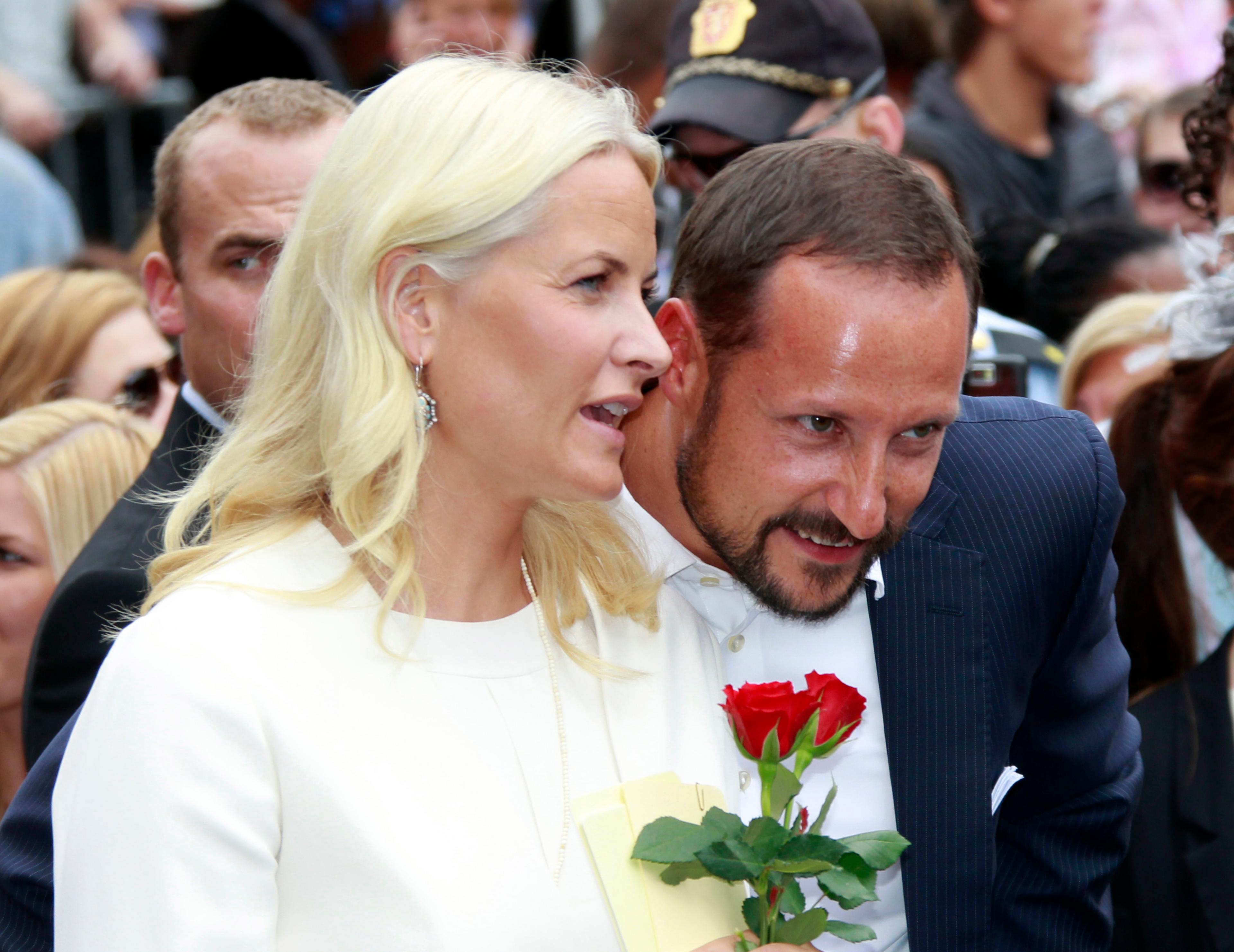 Kronprinsparet blant publikum. Kronprinsessen holder to røde roser og sier noe til Kronprinsen, som lener seg smilende fram for å høre.