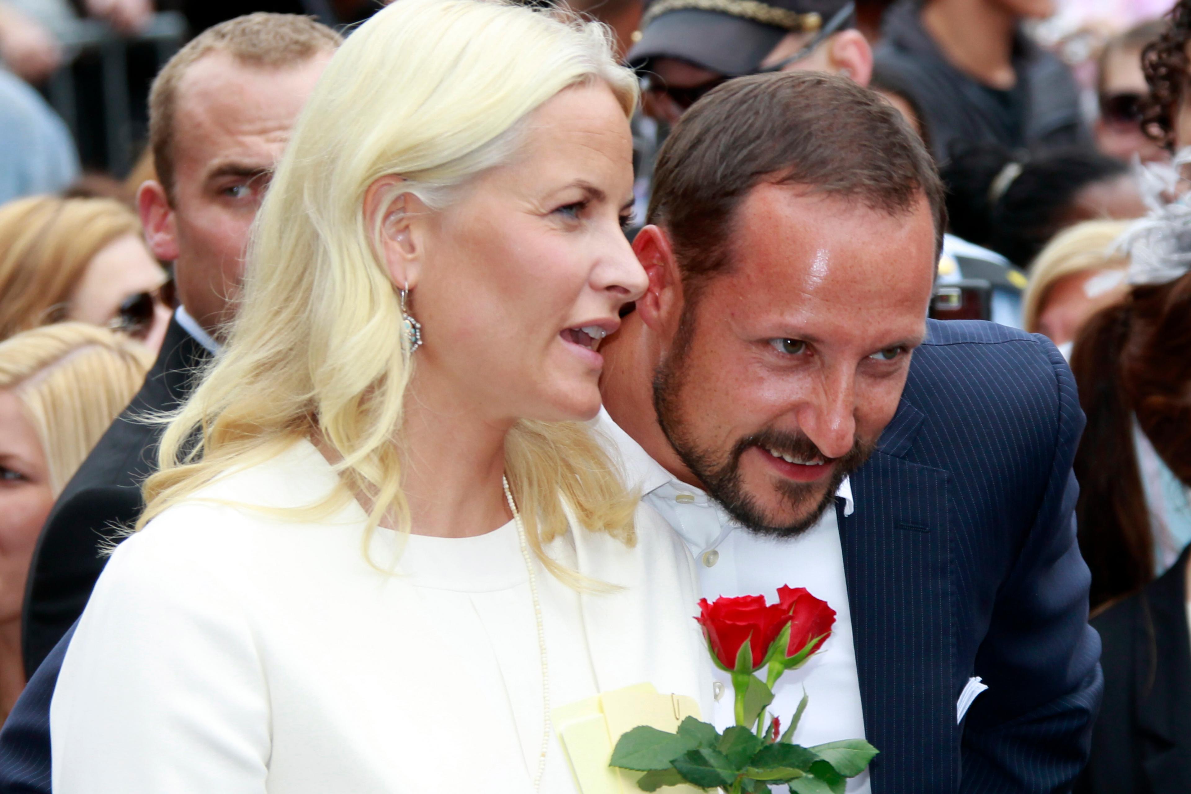 Kronprinsparet blant publikum. Kronprinsessen holder to røde roser og sier noe til Kronprinsen, som lener seg smilende fram for å høre.
