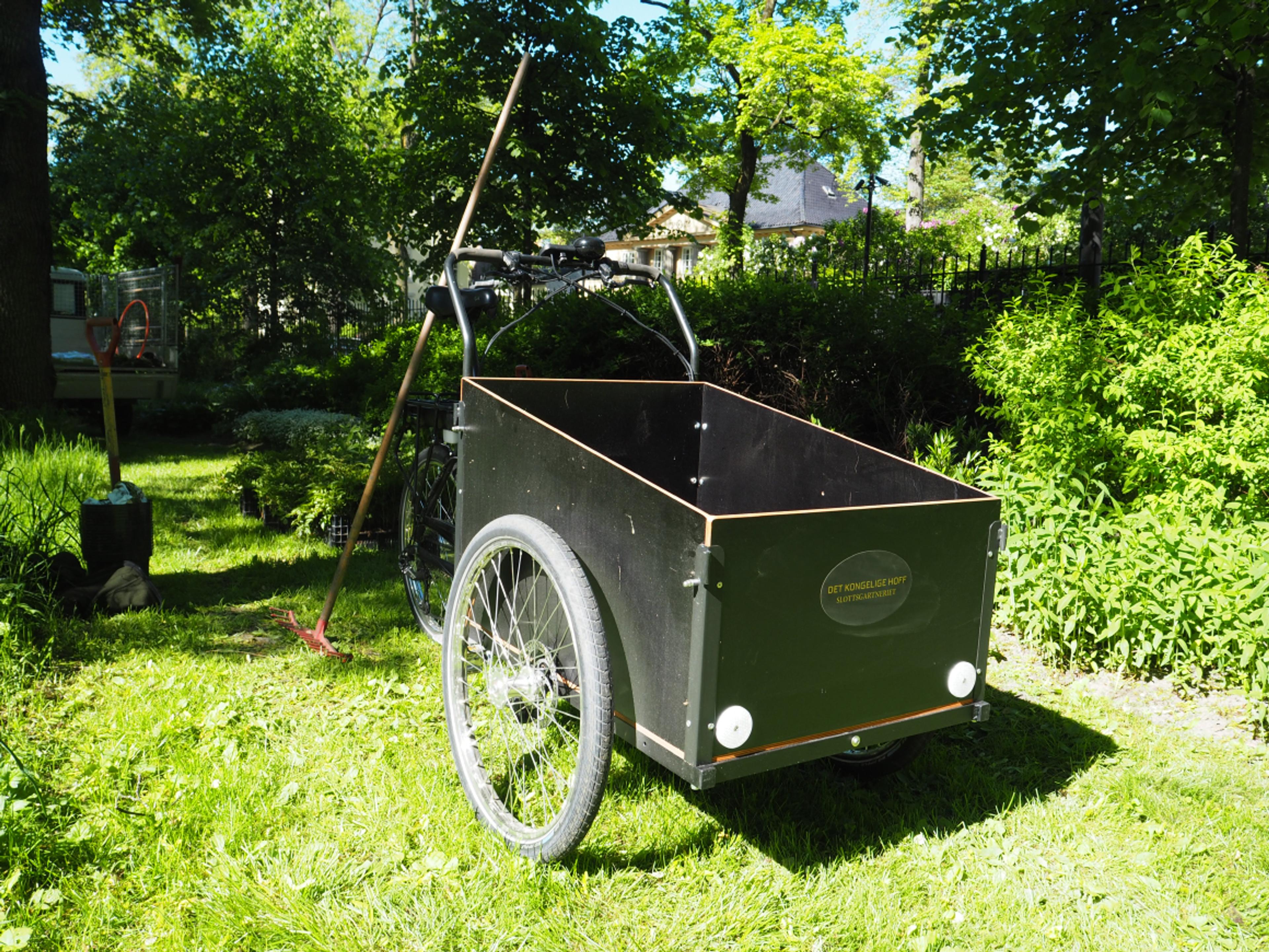 En lastesykkel står ute i parken. Den har en romslig kasse til last og bærer Slottsgartneriets logo.