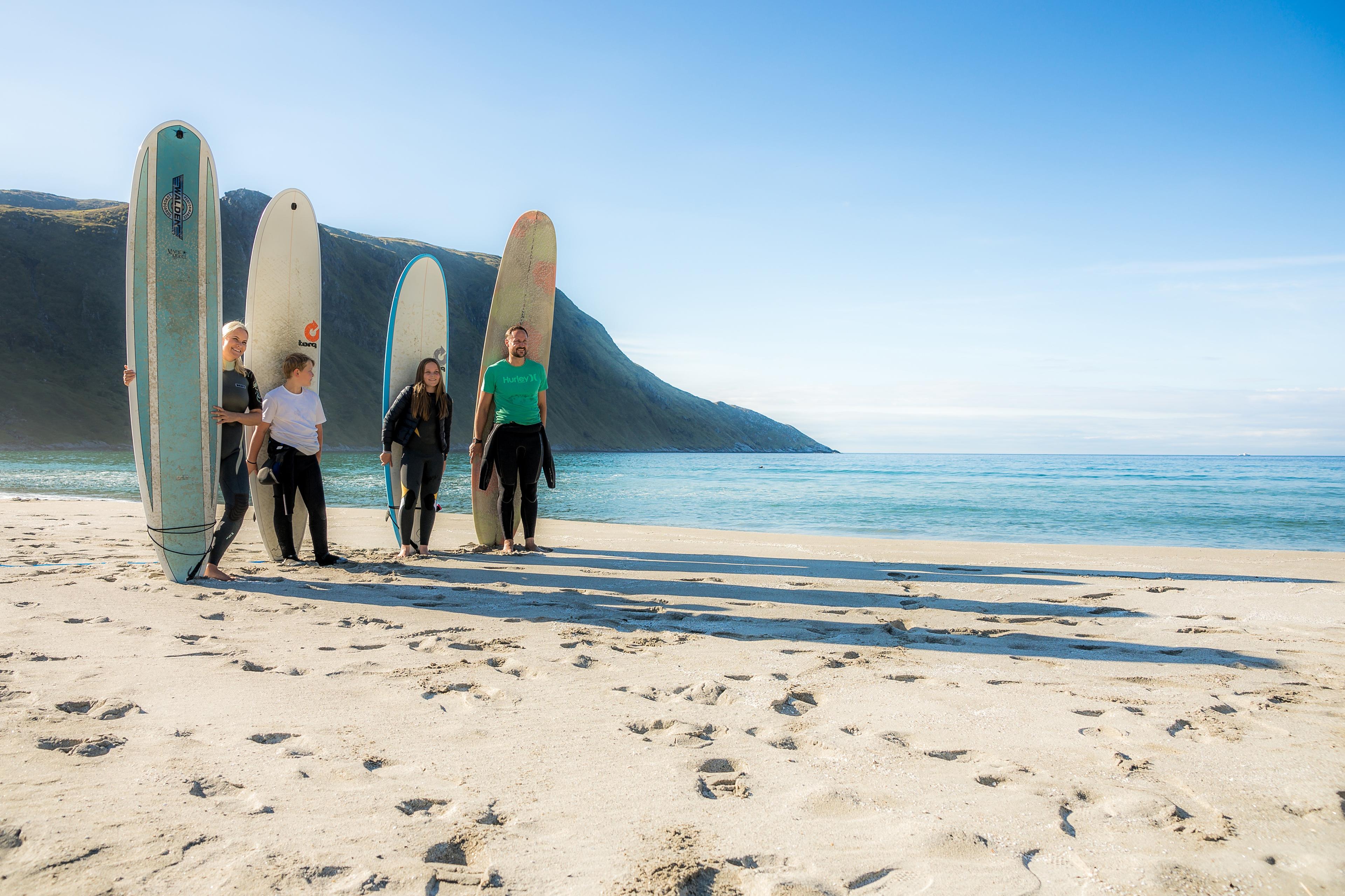Kronprinsparet, Prinsessen og Prinsen står på en sandstrand foran hvert sitt surfebrett og med havet i bakgrunnen.