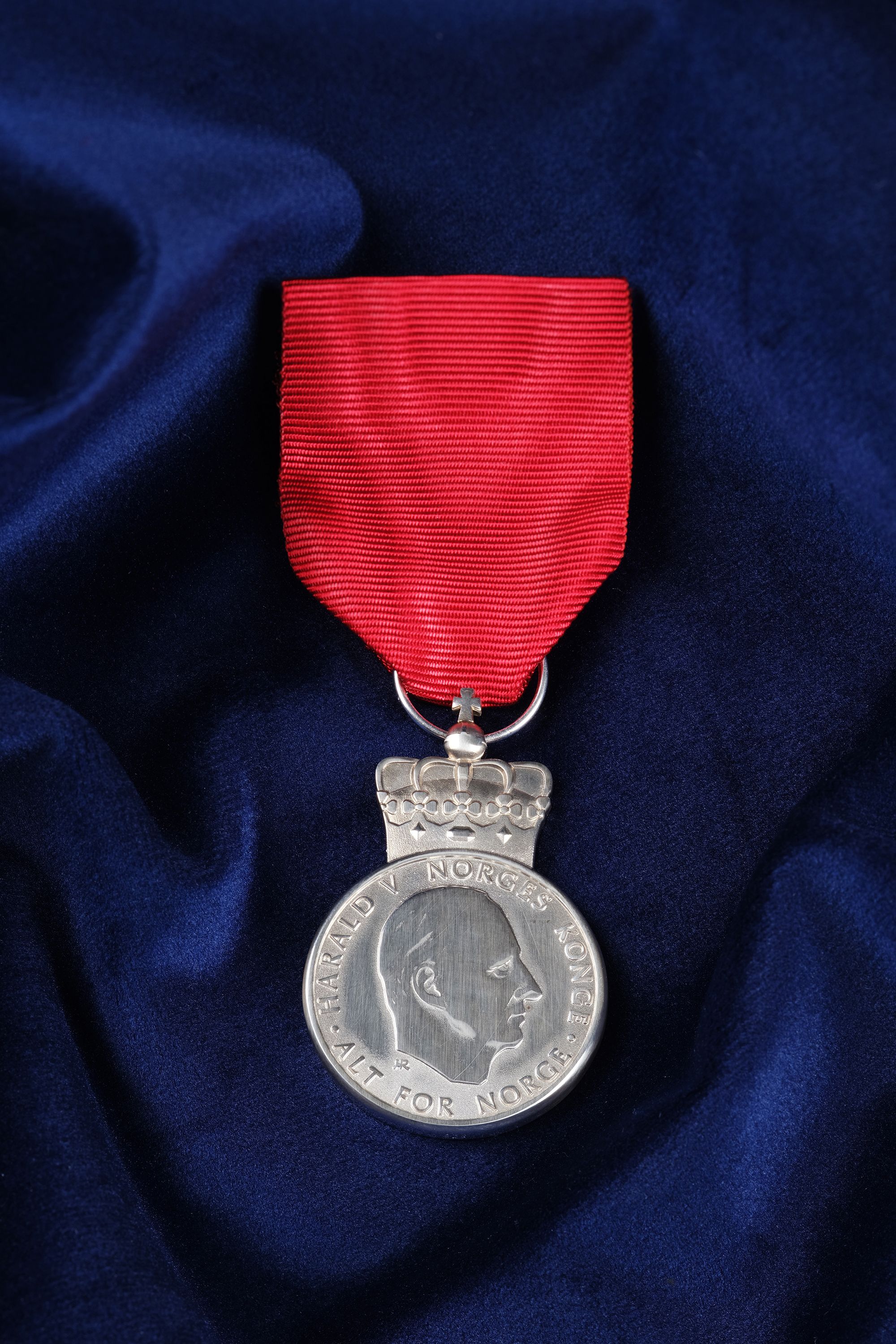 Sølvmedalje preget med Kongens portrett, festet i et rødt bånd. Ligger på på et dypblått klede.