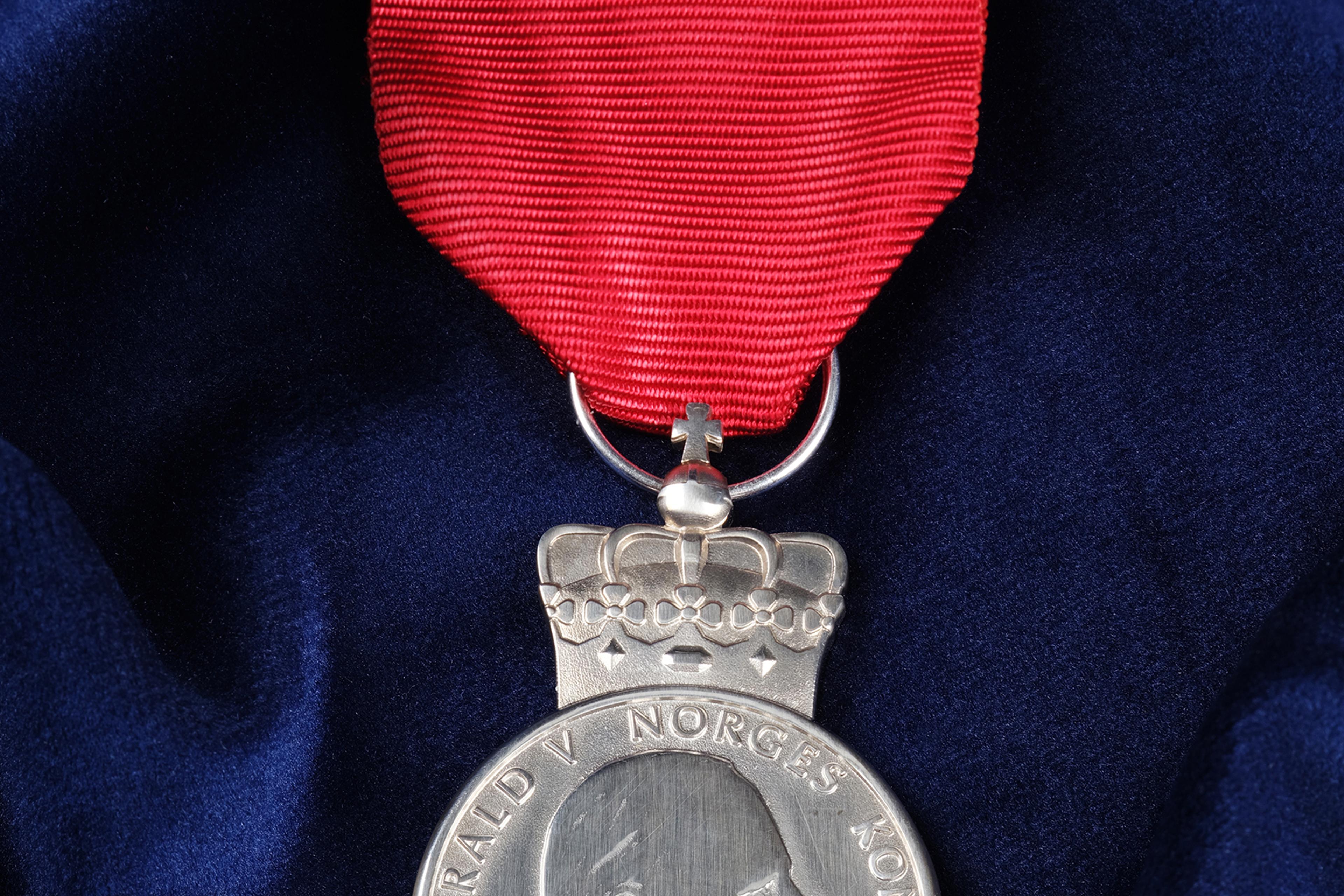 Sølvmedalje preget med Kongens portrett, festet i et rødt bånd. Ligger på på et dypblått klede.