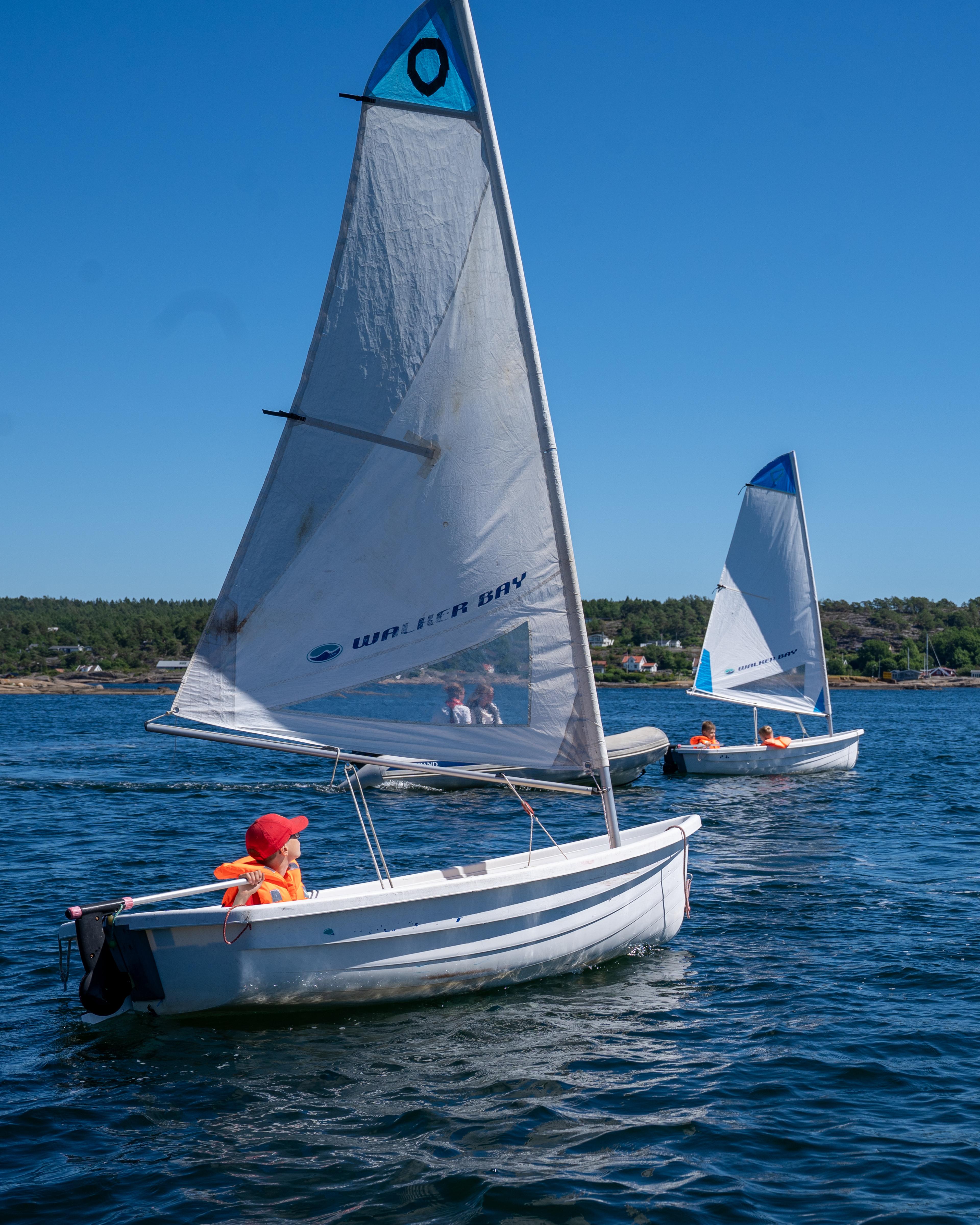 En ung gutt med redningsvest sitter i en liten seiljolle på vannet.