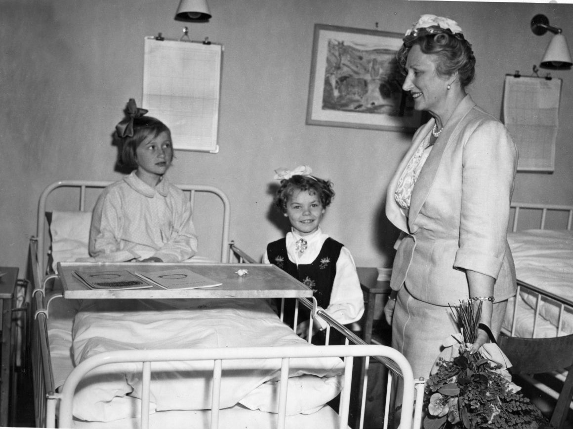 Kronprinsessen står ved siden av en sykeseng med en liten pike i.