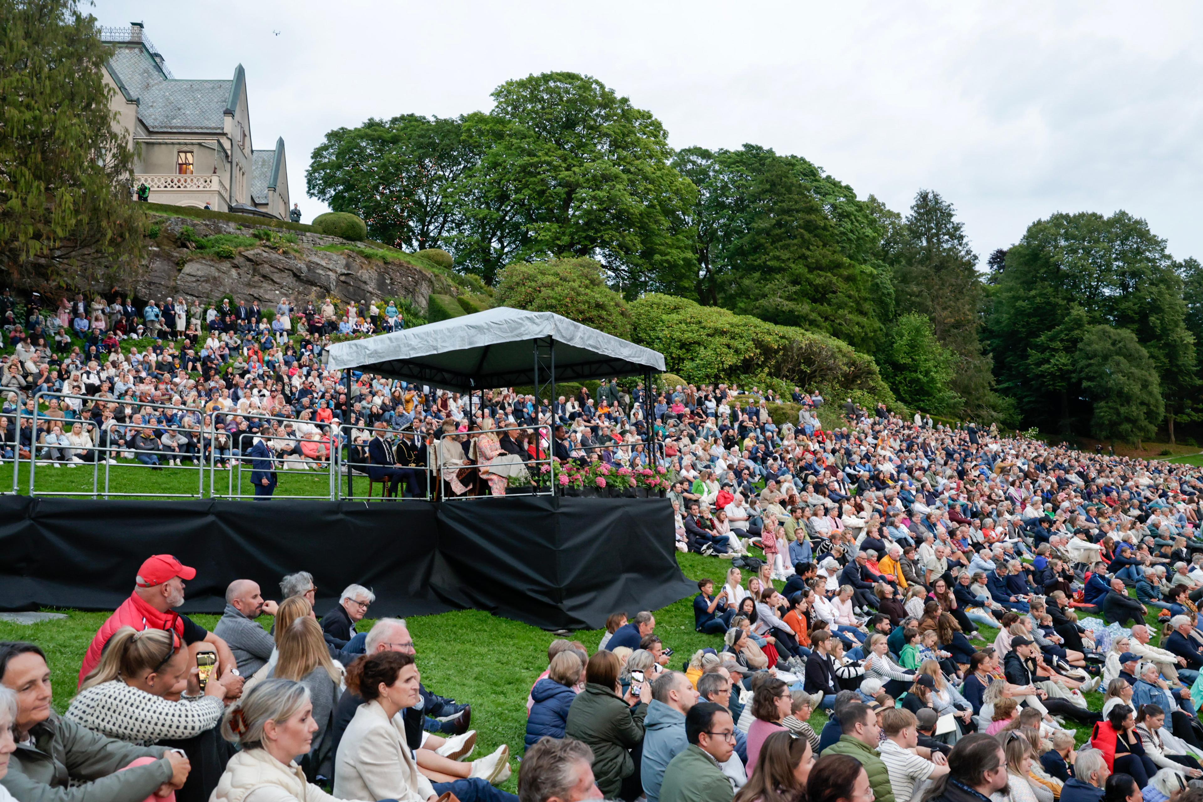 Mengder av mennesker sitter på bakken i et naturlig atrium og ser mot scenen utenfor bildet. De kongelige sitter i en paviljong i sentrum av bildet og bak ses en del av herskapshuset.