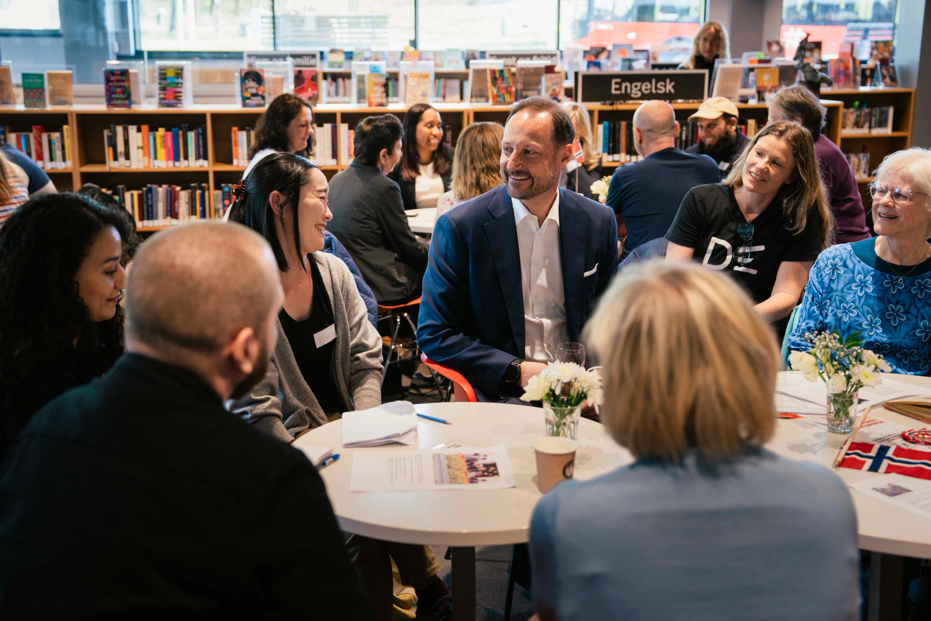 Kronprins Haakon sitter i en gruppe med mennesker rundt et bord på inne på et bibliotek, med bøker i bakgrunnen.