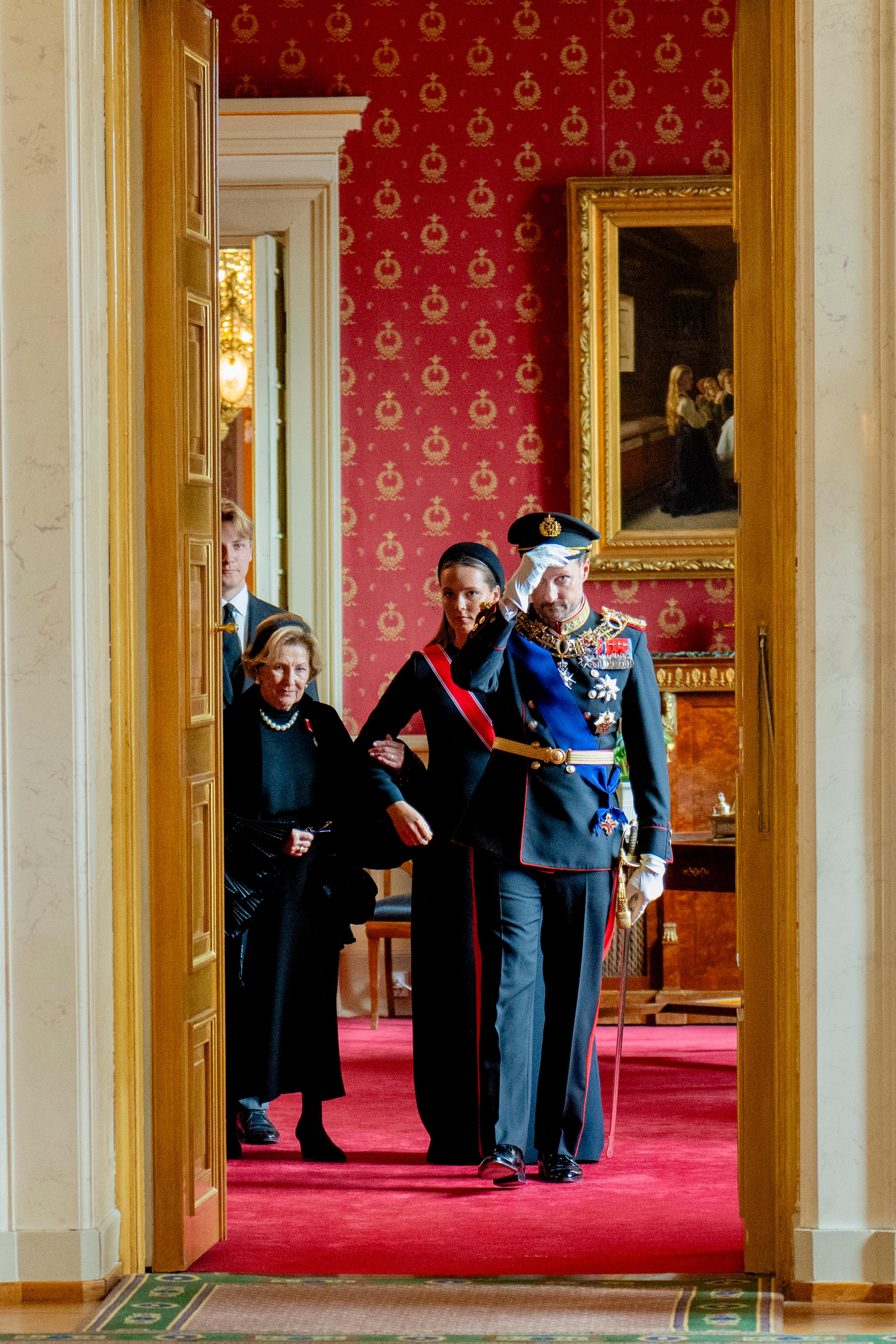 Sett gjennom den åpne døren til Røde salong: Kong Haakon setter uniformsluen på hodet og begynner å gå. Bak ham kommer Kronprinsessen og Dronning Sonja.