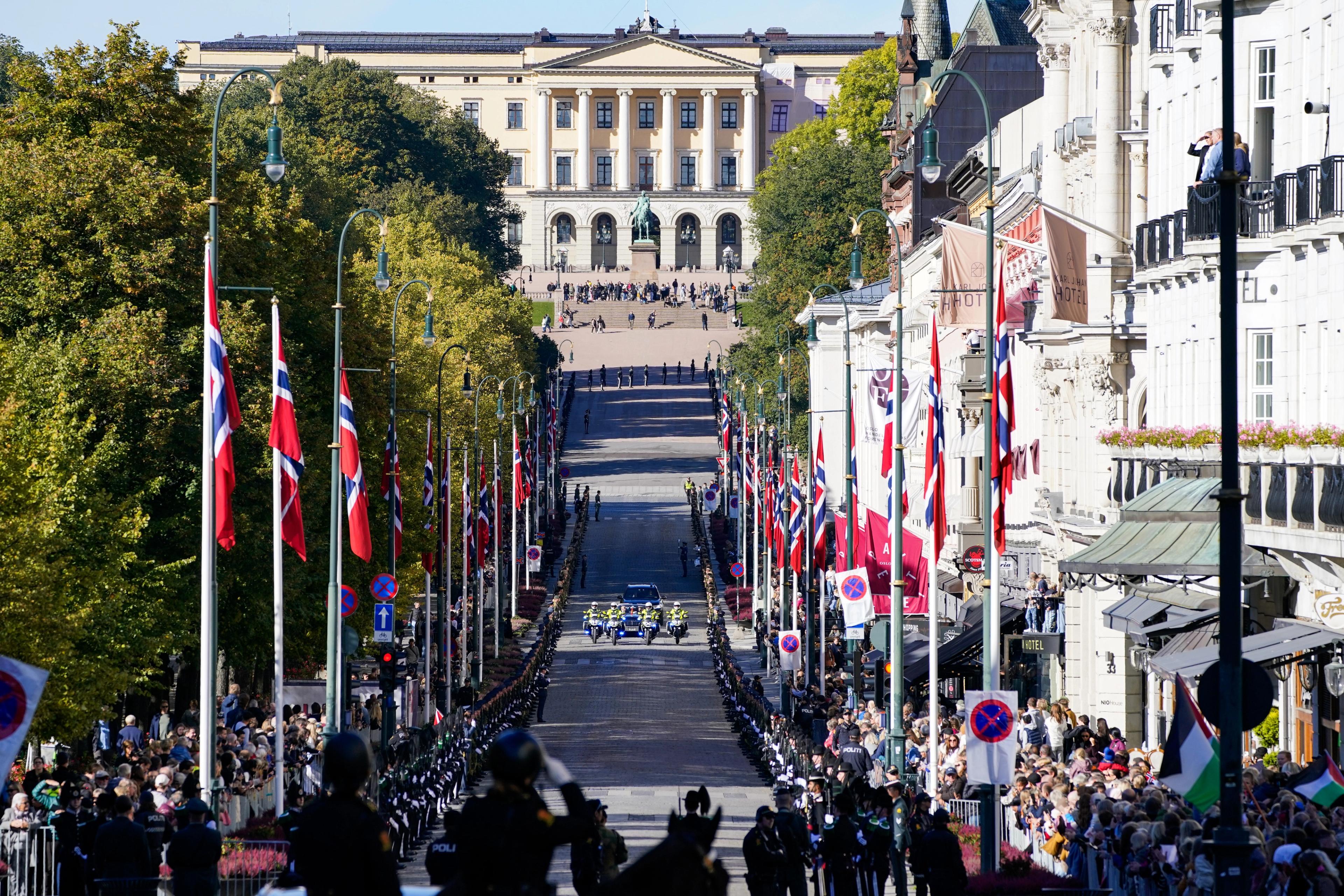 Fotografen har stått midt i gaten og fotografert oppover mot Slottet. Det vaier norske flagg på begge sider av gaten og en bilkortesje nærmer seg.