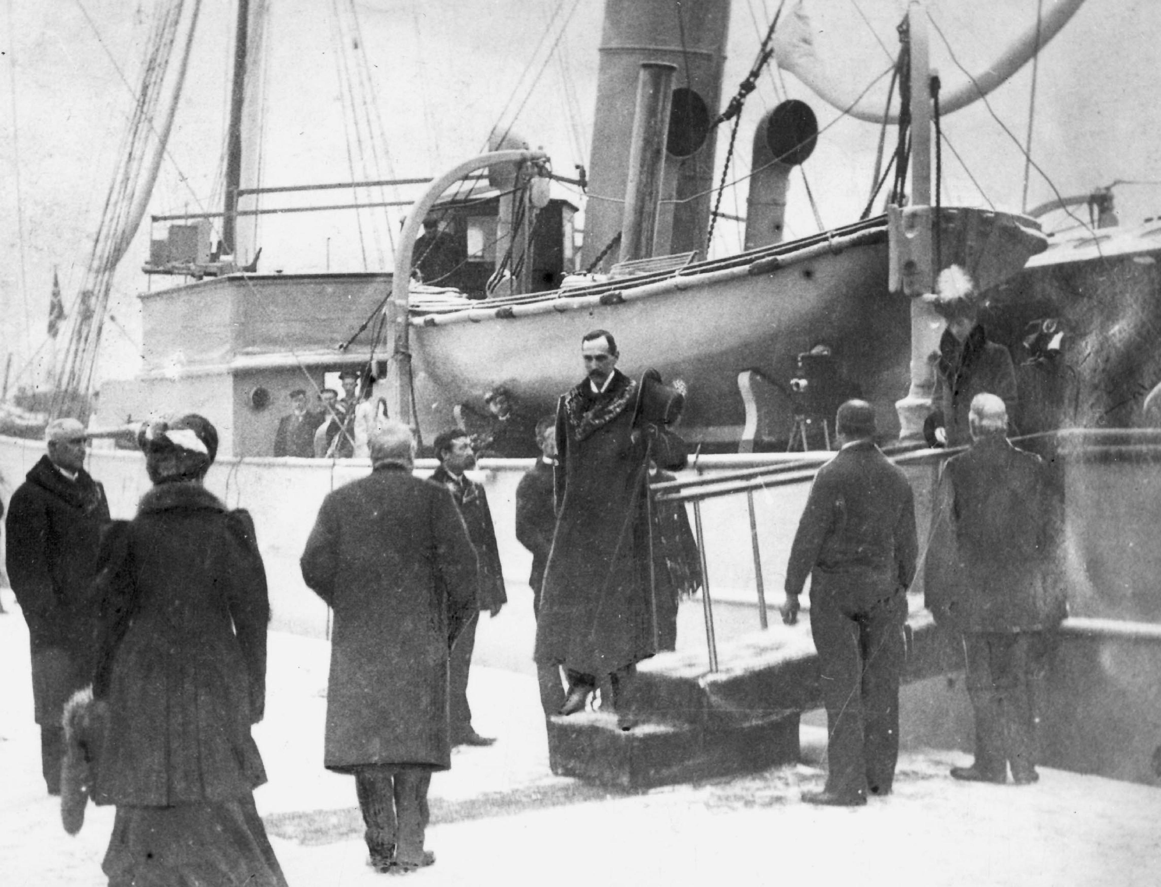 Kong Haakon står på tuppen av gangveien, i ferd med å sette foten på norsk jord. I land står en mottakelseskomité og venter. Kongen har tatt av seg hatten for å hilse.