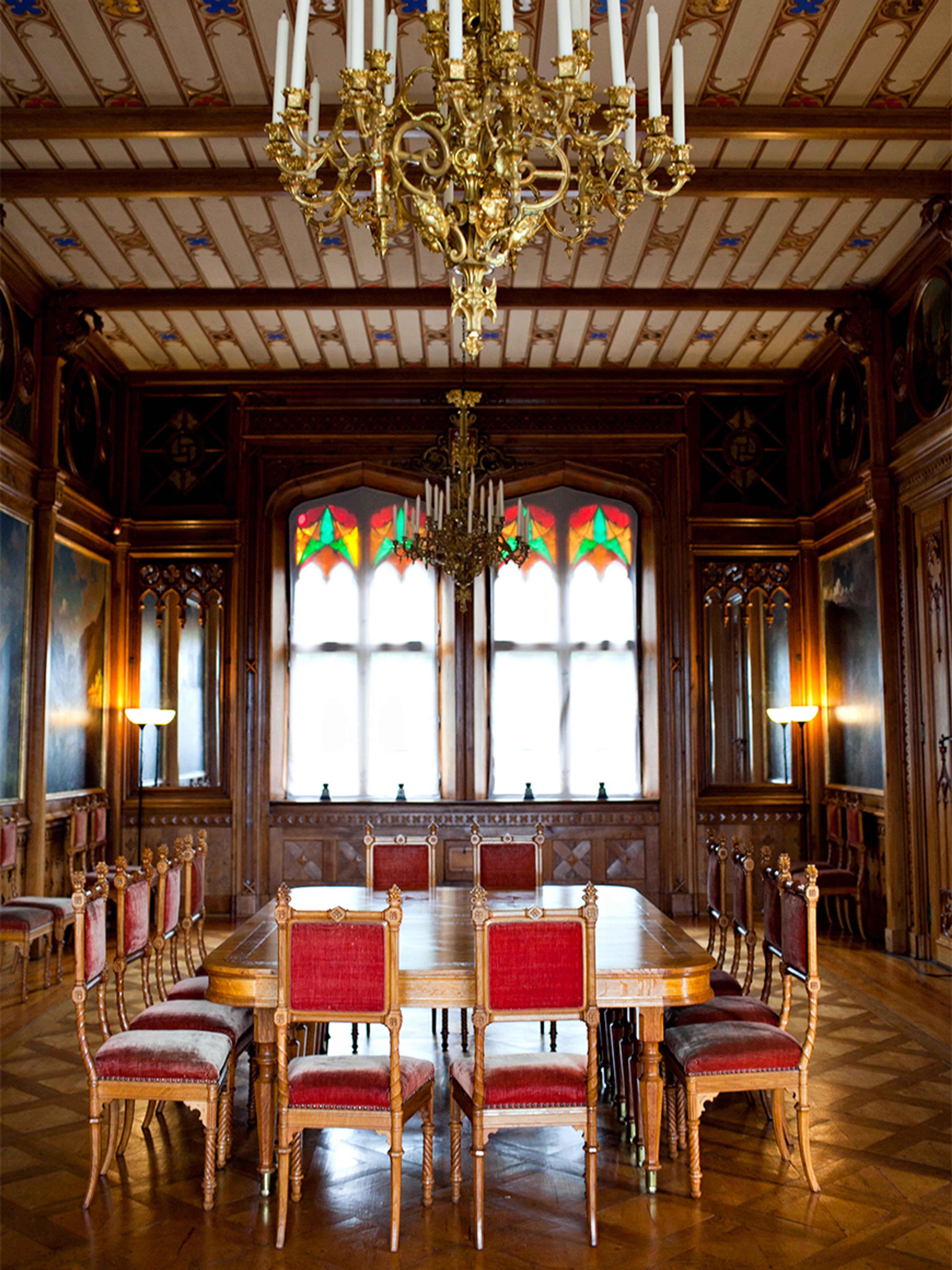 Bord og stoler står midt i et rom med store vinduer og vegger utsmykket med malerier.