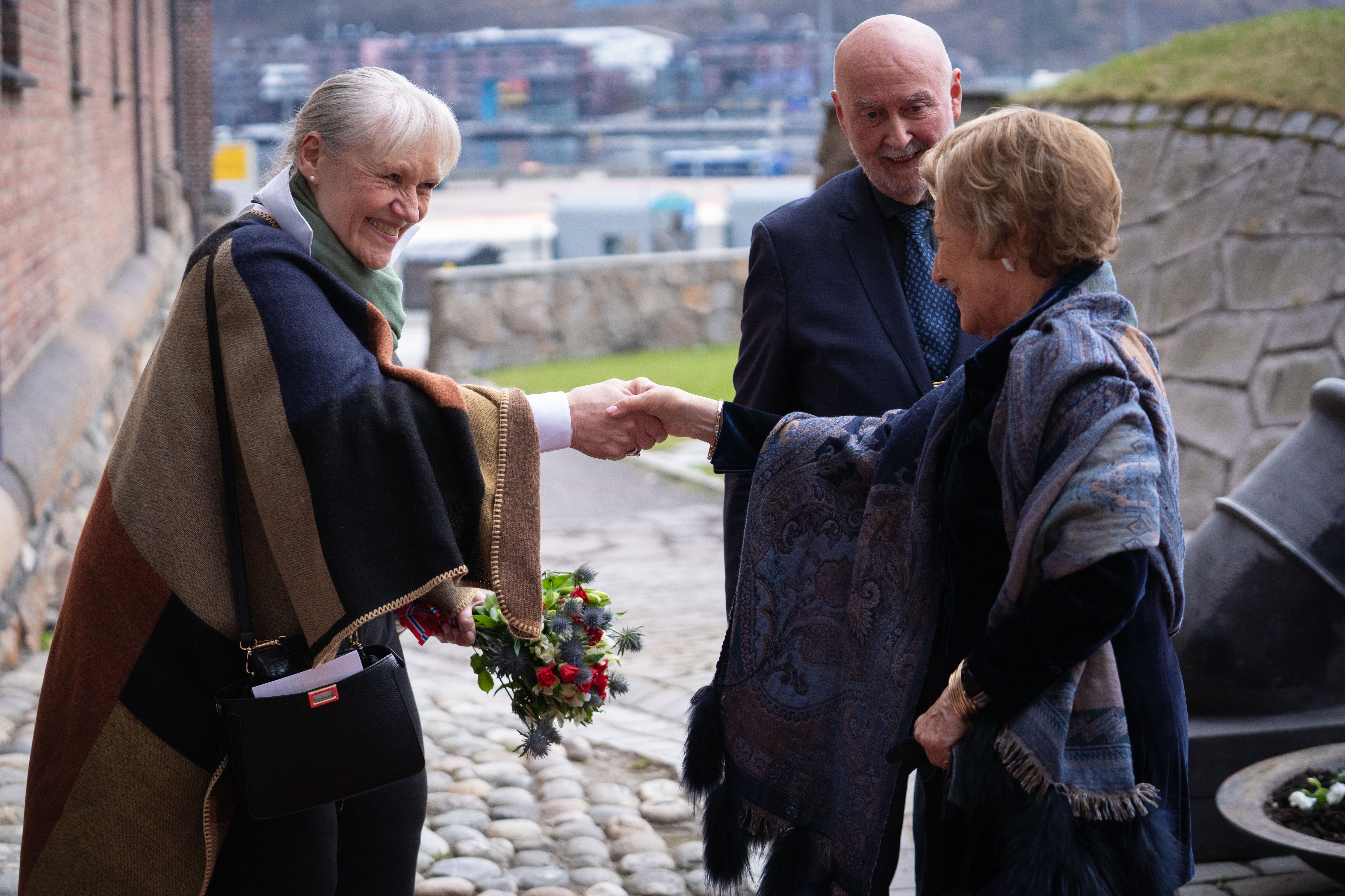 Daglig leder hilser på Dronning Sonja utendørs på Akershus festning. Styreleder står i bakgrunnen.