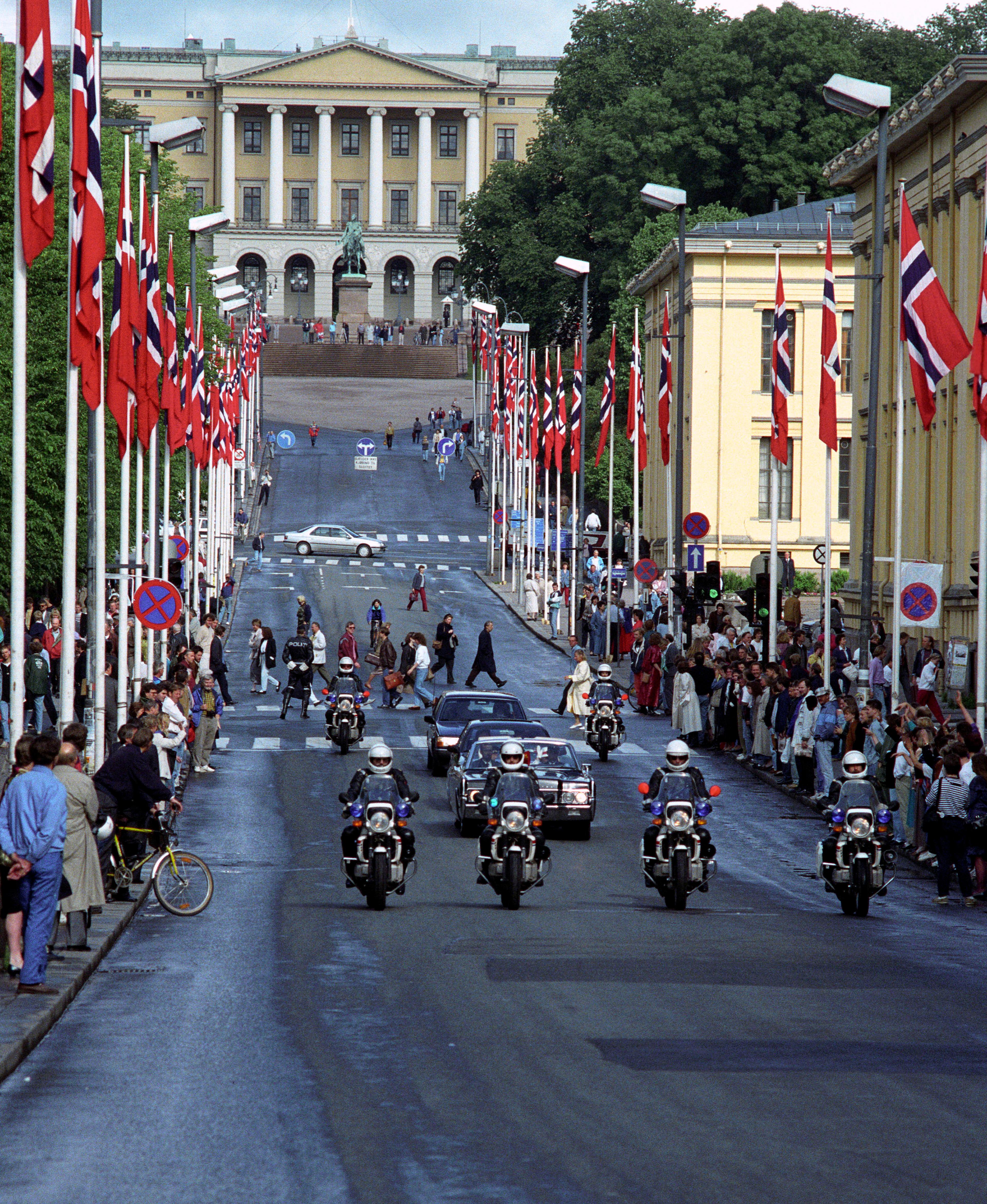 Bilkortesje på vei ned Karl Johans gate. Fire motorsykler foran, så to biler og ytterligere to motorsykler. Norske flagg vaier på begge sider av gaten. Slottet danner bakgrunnen i bildet.