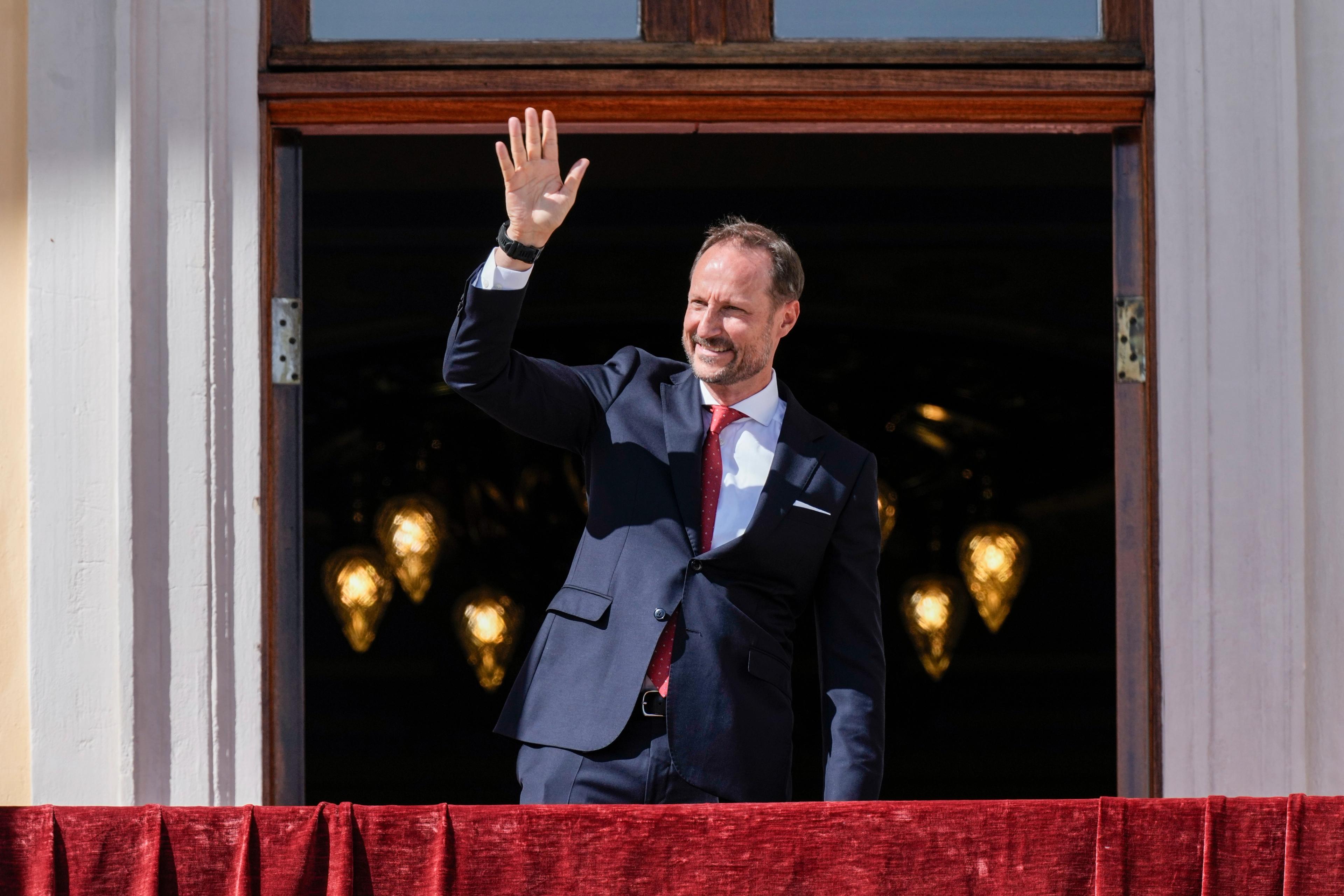 Kronprins Haakon vinker fra Slottsbalkongen ikledd dress.