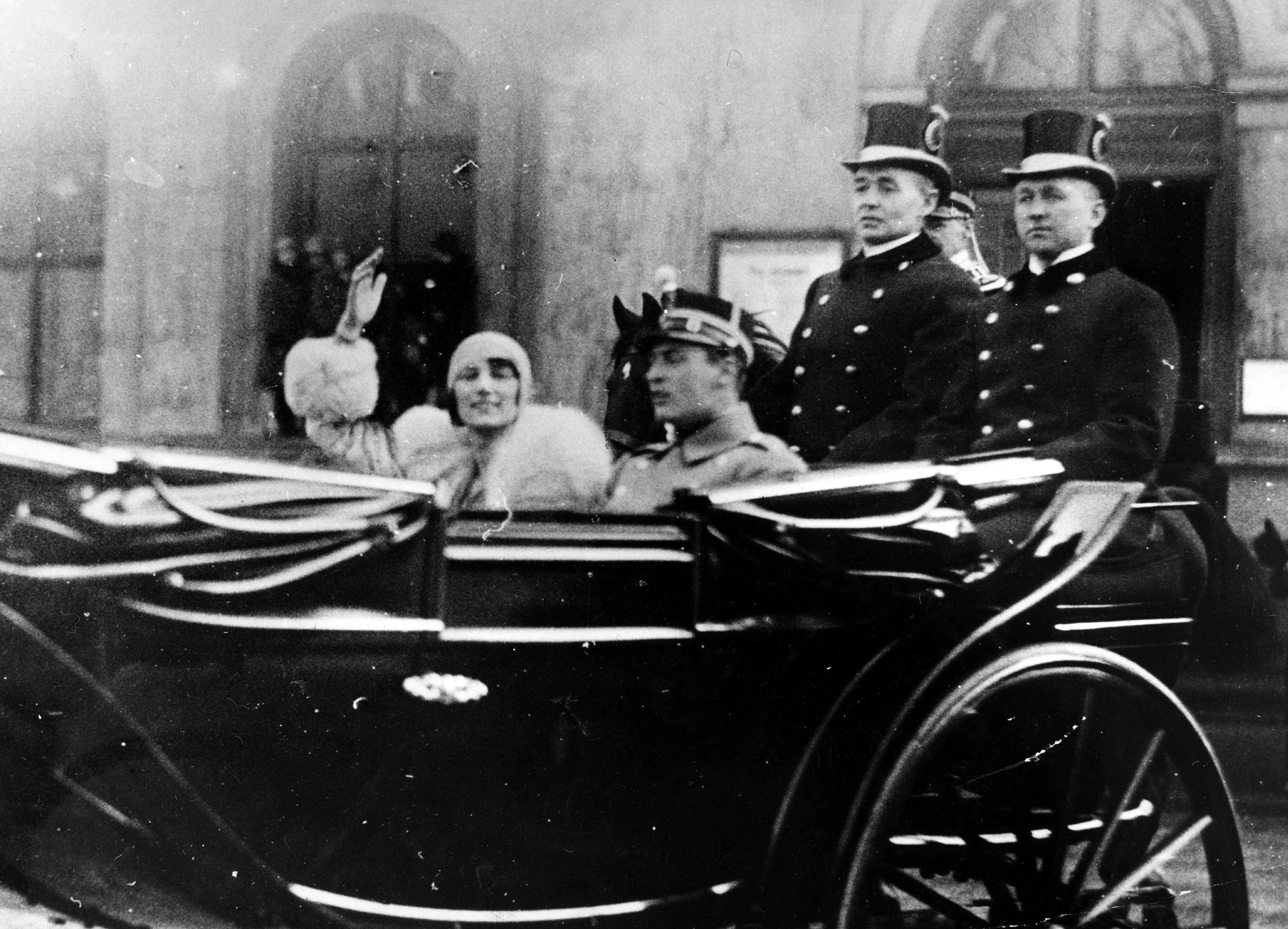 Kronpins Olav og Prinsesse Märtha sitter i en åpen vogn. Prinsessen vinker.