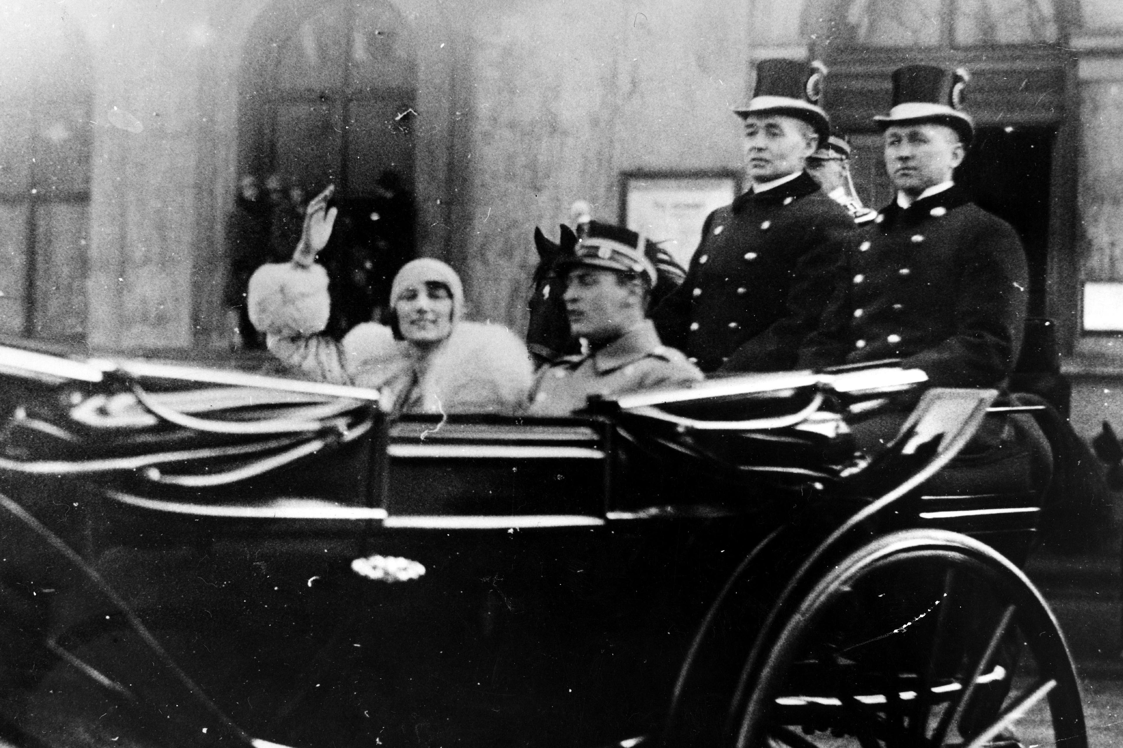 Kronpins Olav og Prinsesse Märtha sitter i en åpen vogn. Prinsessen vinker.