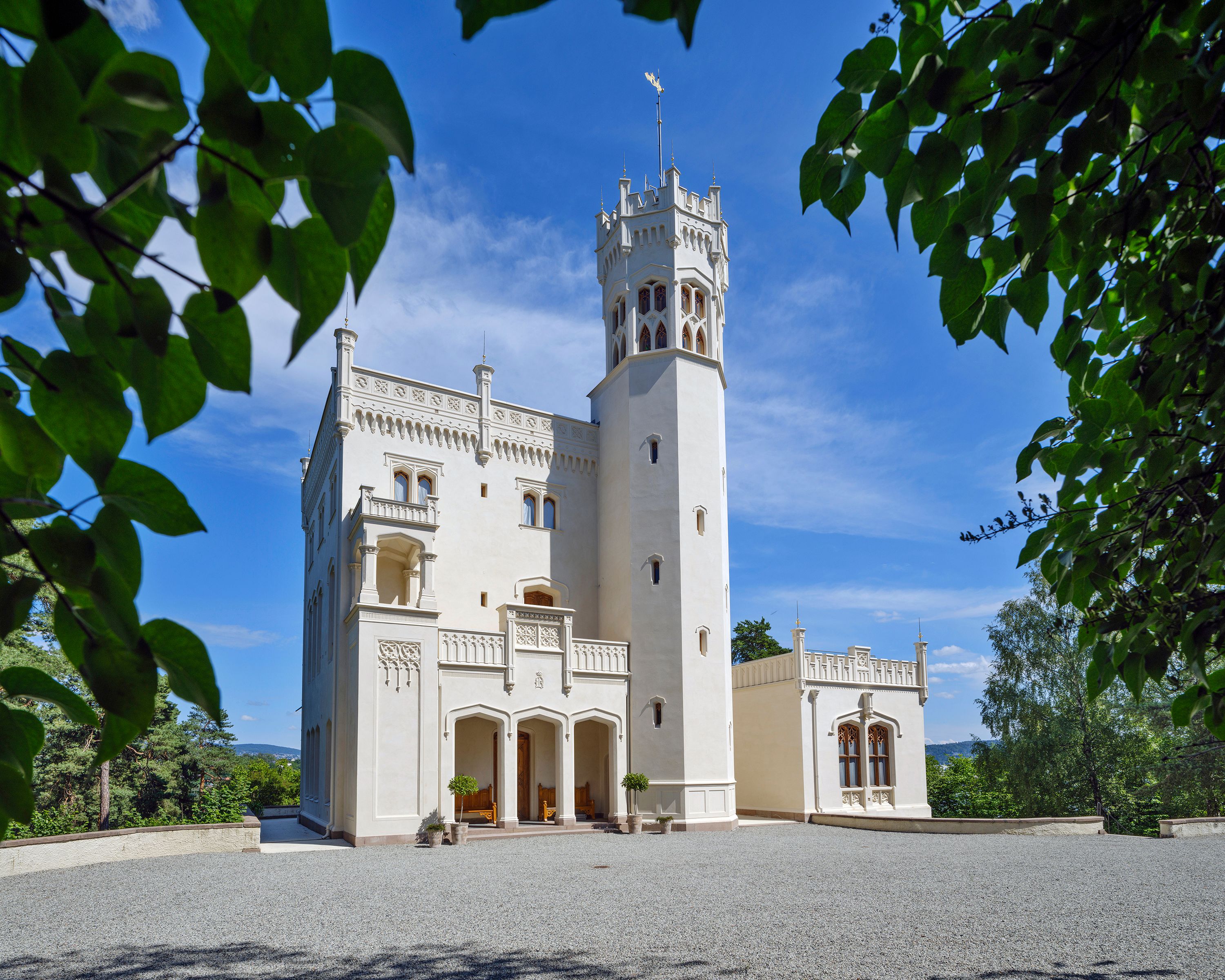 Oscarshalls fasade sett gjennom omkransende bladverk. Det lille slottet er hvitt og har både svalgang og tårn.