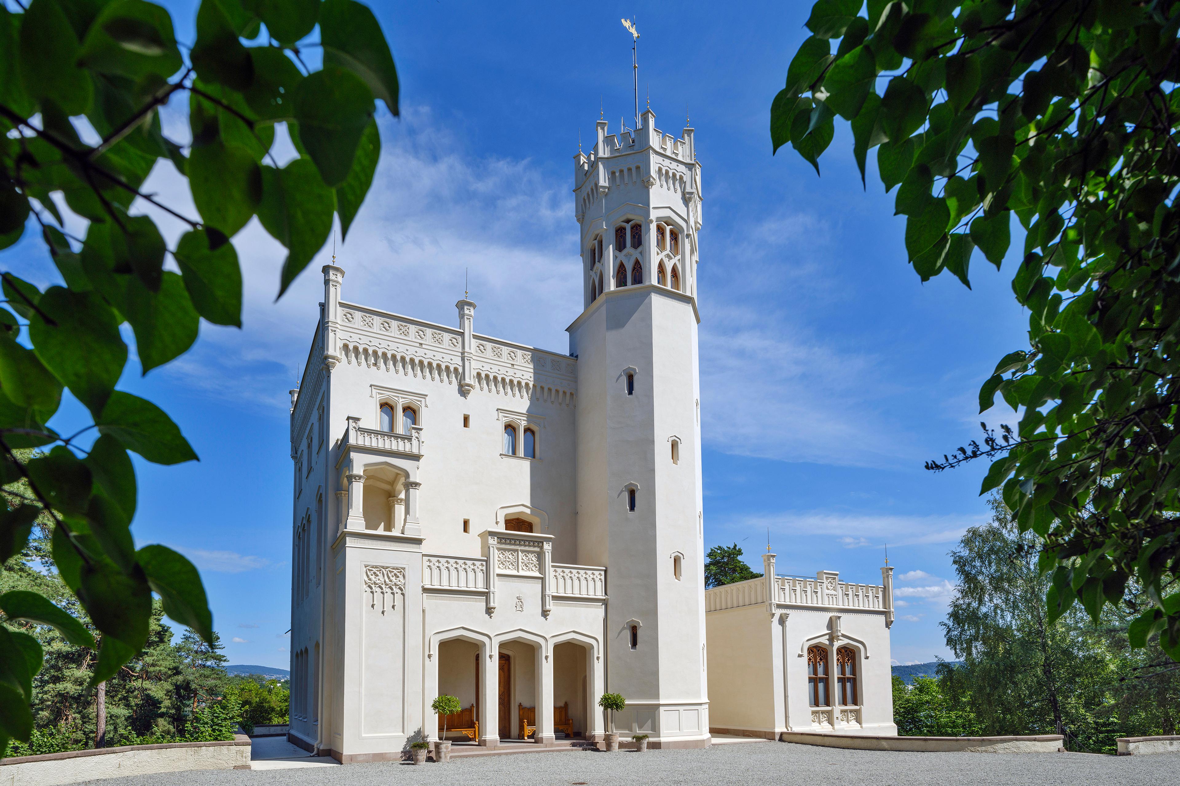 Oscarshalls fasade sett gjennom omkransende bladverk. Det lille slottet er hvitt og har både svalgang og tårn.