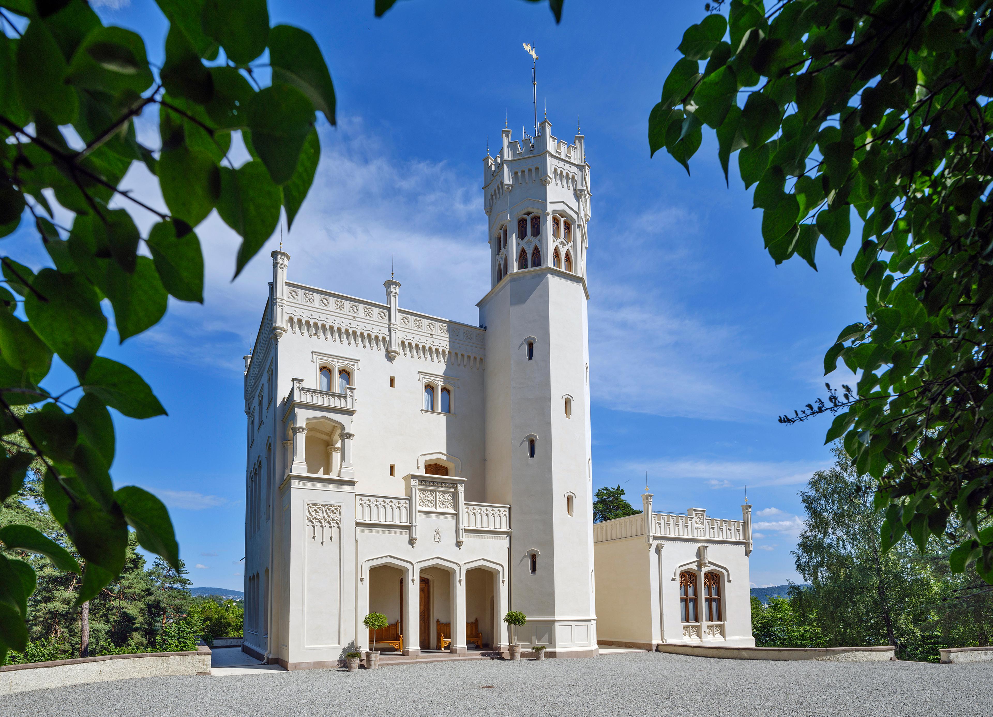 Oscarshalls fasade sett gjennom omkransende bladverk. Det lille slottet er hvitt og har både svalgang og tårn.