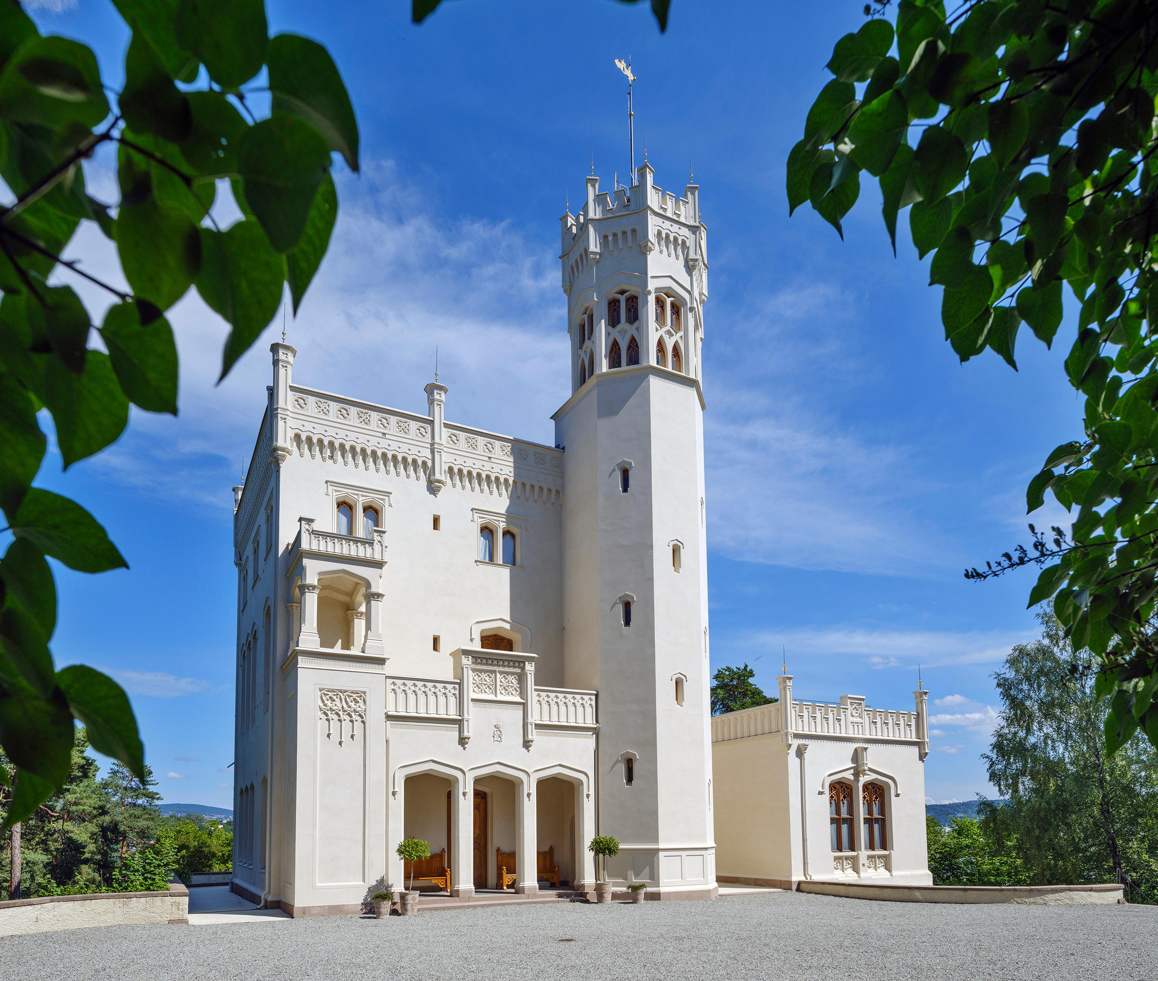 Oscarshalls fasade sett gjennom omkransende bladverk. Det lille slottet er hvitt og har både svalgang og tårn.