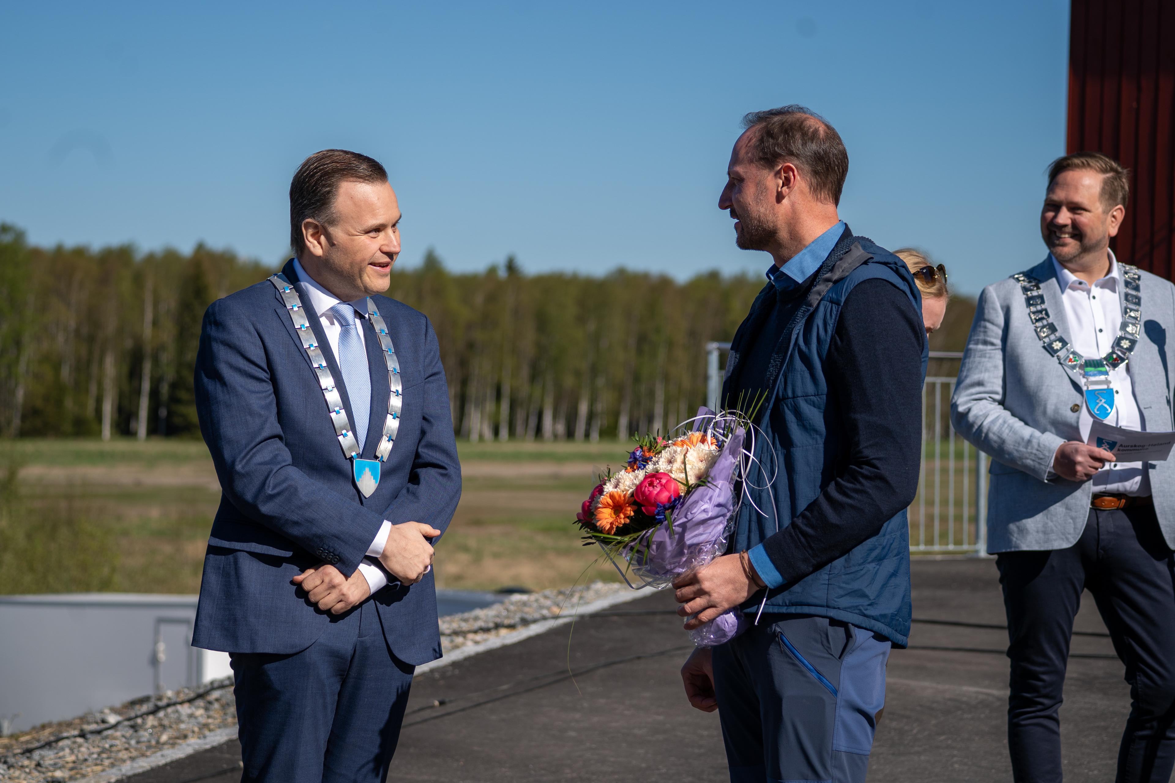 Kronprins Haakon står med blomstebukett i hånden, i samtale med fylkesordføreren med fylkeskjede rundt halsen.