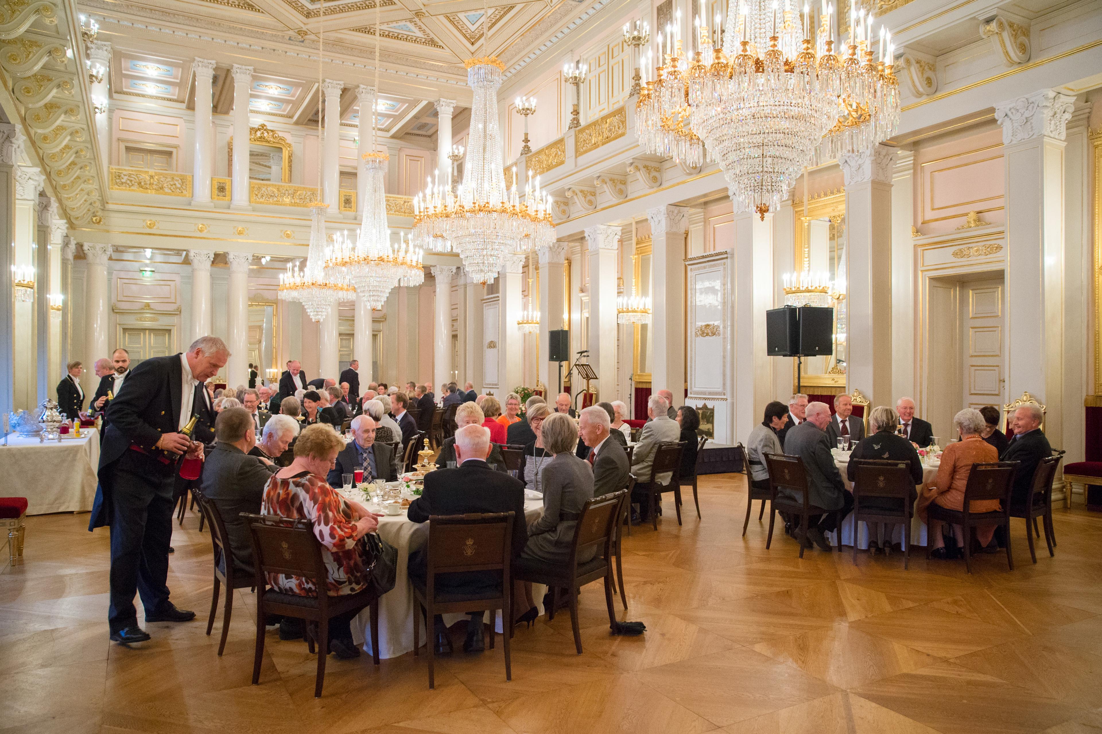 Pyntede mennesker sitter ved runde bord i et stort rom dekorert i hvitt og gull. Tre store lysekroner henger ned fra taket. En livrékledd servitør byr gjestene på drikke.