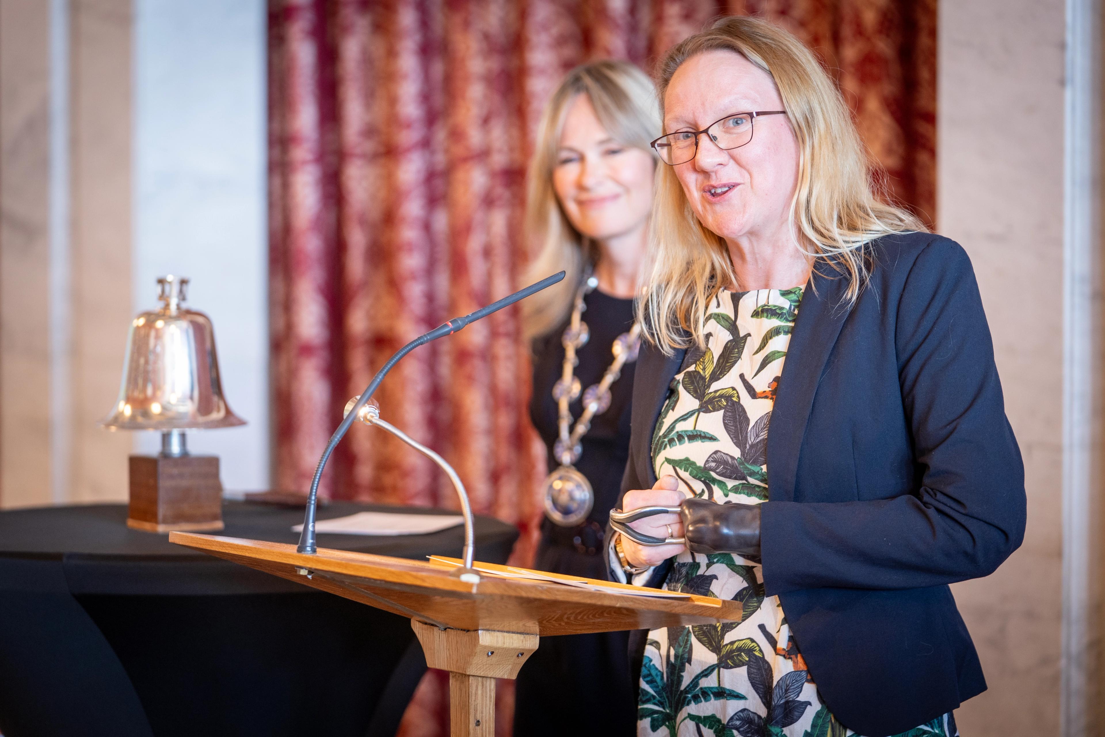 Caroline Solem står på en talerstol. I bakgrunnen står ordfører Anne Lindboe.