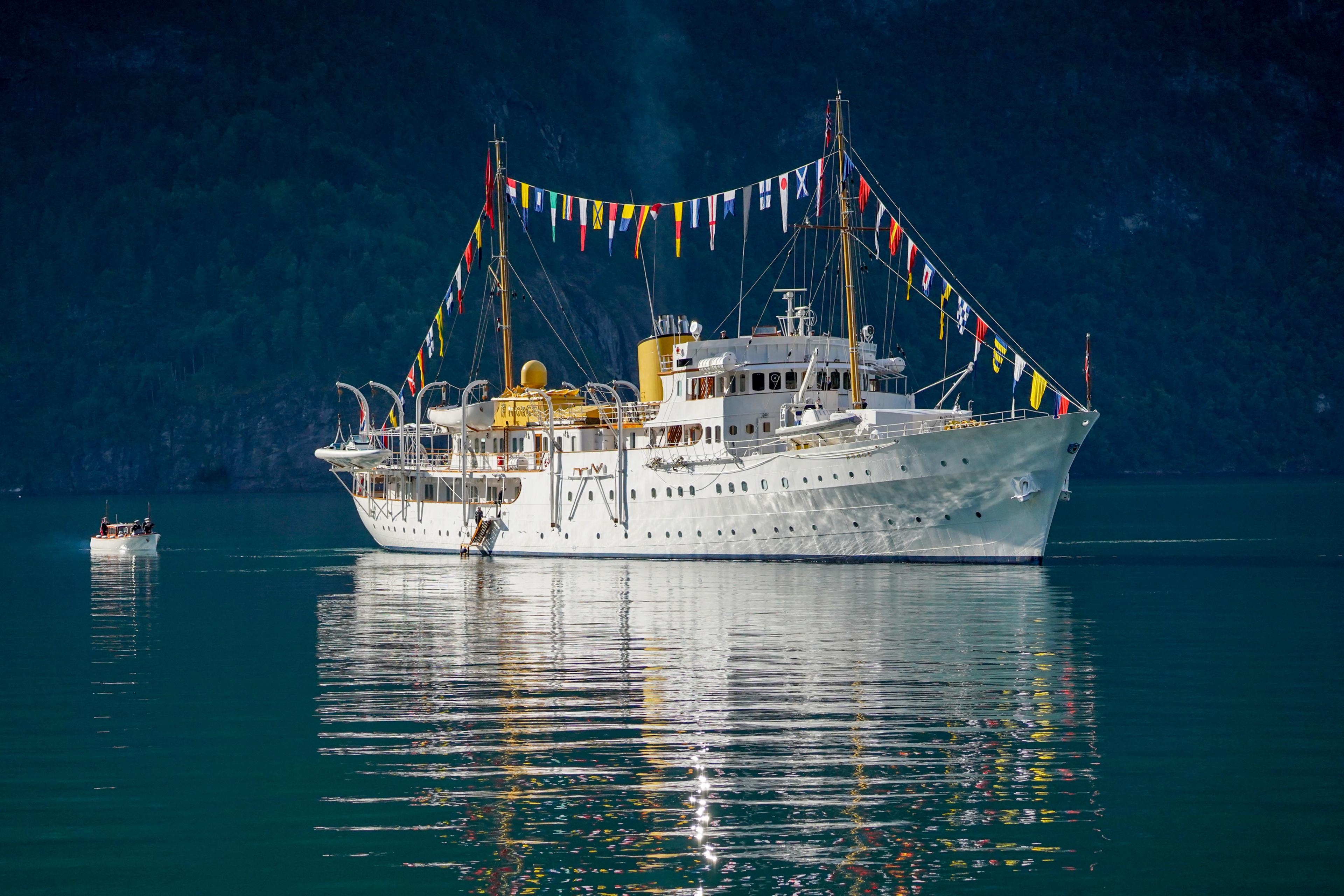 Kongeskipet fra siden med festflagg mellom mastene. Speiler seg i blankt hav.