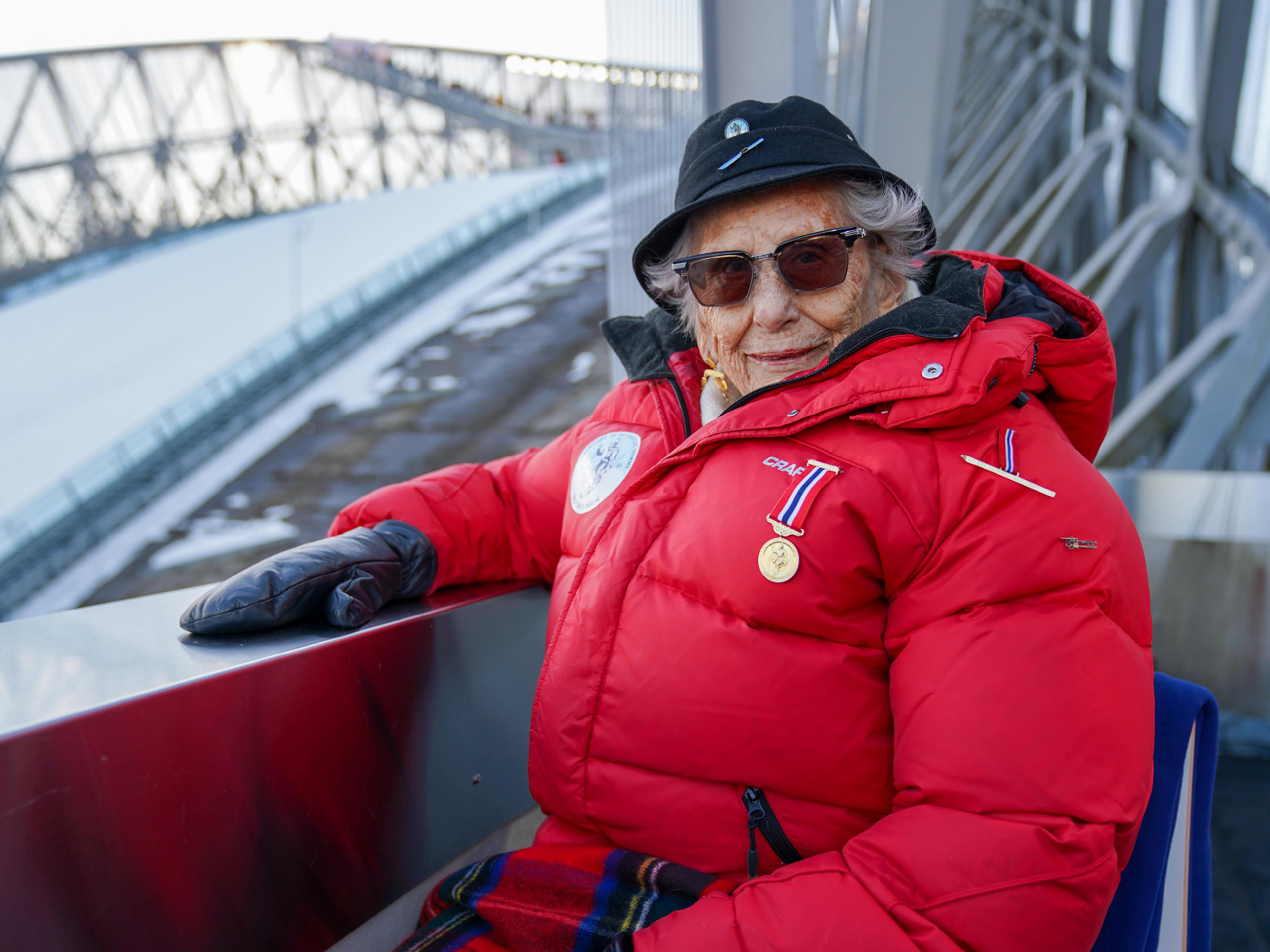 Prinsesse Astrid på kongetribunen i Holmenkollen, godt kledd for vinterværet, med hoppet i bakgrunnen og Holmenkollmedaljen festet til den røde jakken.