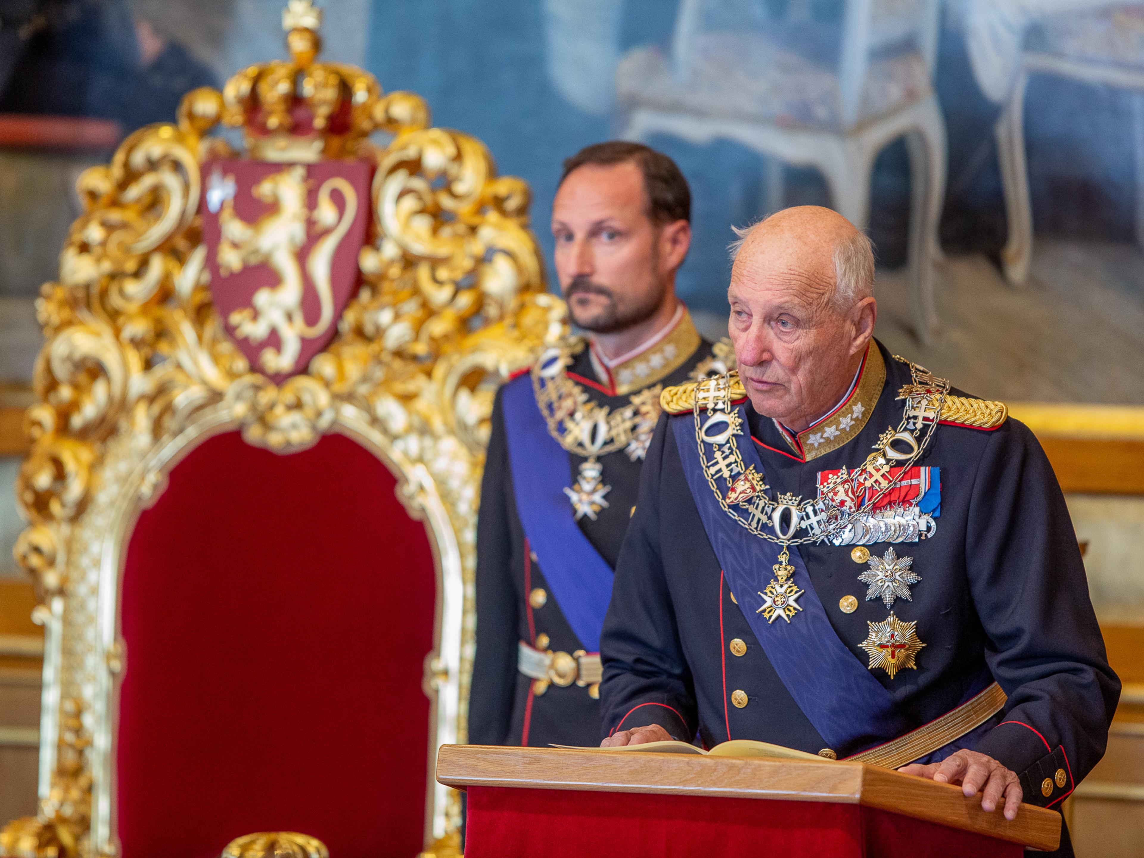 Kong Harald står i gallauniform på Stortingets talerstol, med Kronprins Haakon i bakgrunnen, ved siden av tronstolen i rødt og gull.
