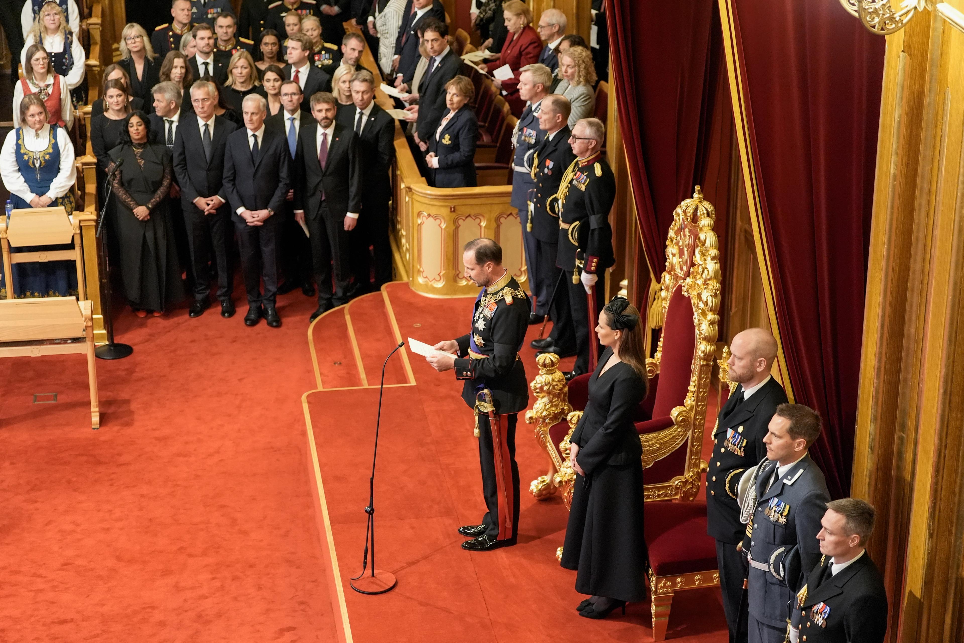 Kong Haakon ser ned på trontalen. Kronprinsesse Ingrid Alexandra står ved hans venstre side. Regjeringen står oppreist på hans høyre side.