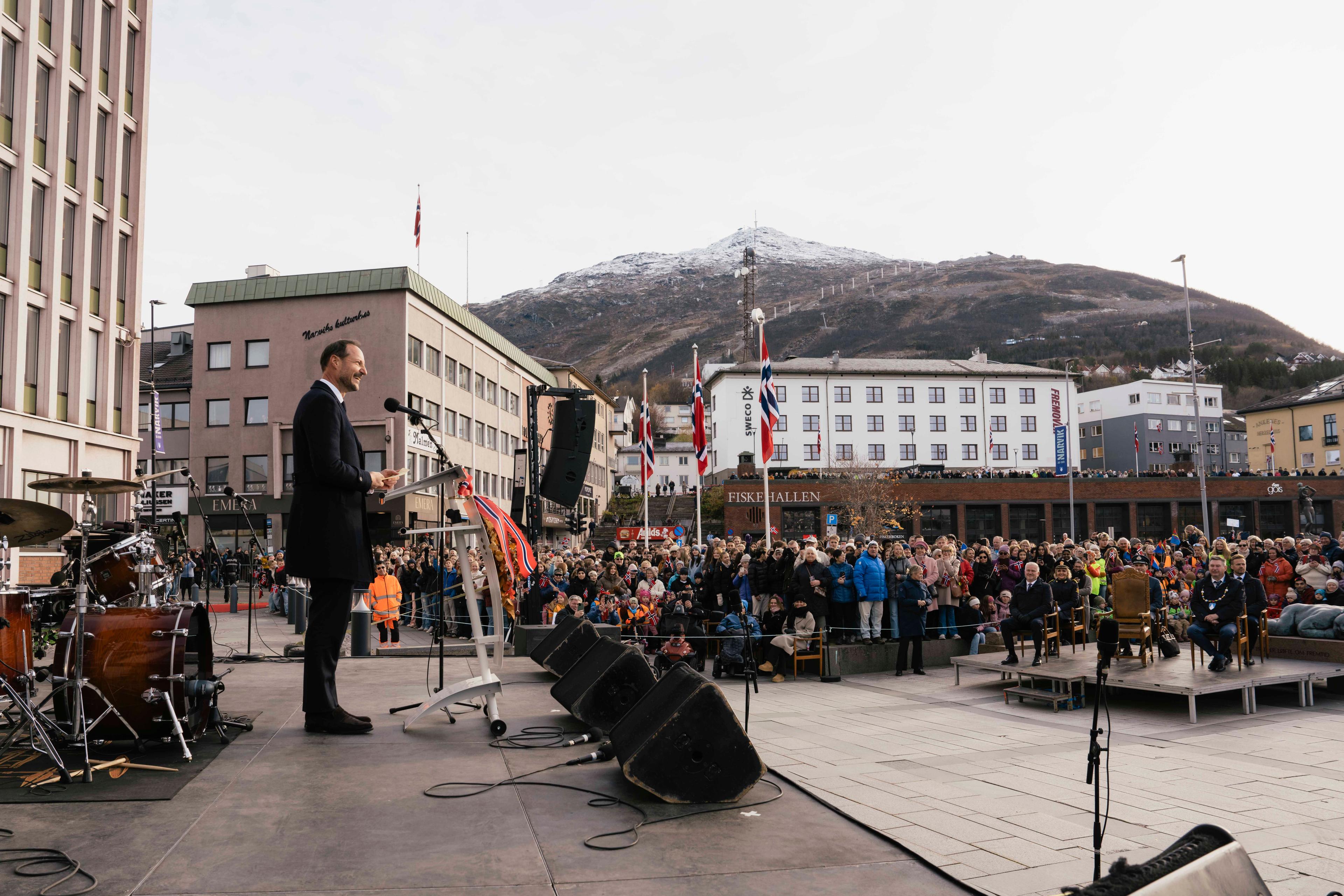 Kong Haakon står på en scene og taler ut til en større folkemenge. Byen Narvik ses i bakgrunnen, med bygg og fjell