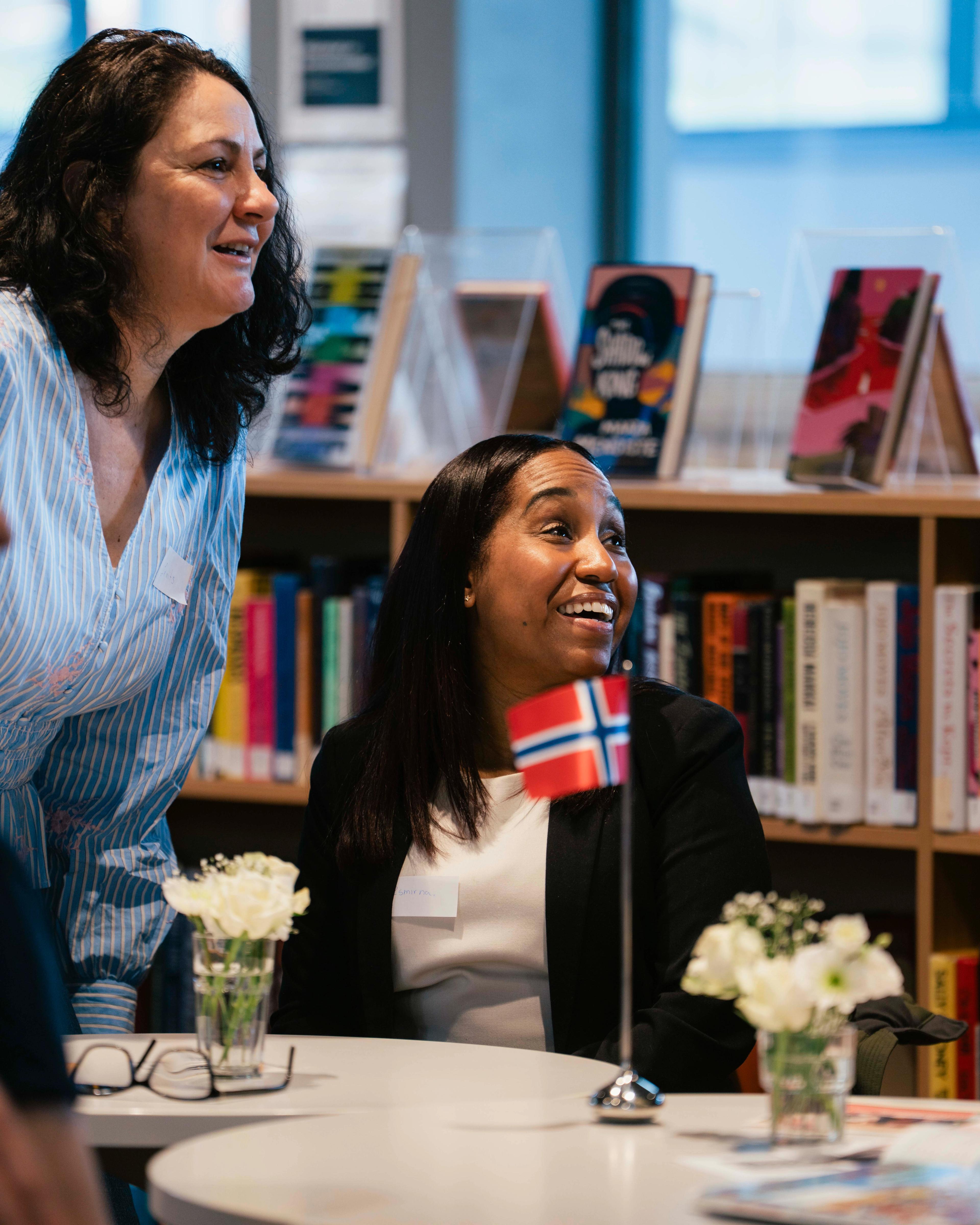 To smilende kvinner rundt bordet på biblioteket. I forgrunnen ser vi et norsk flagg