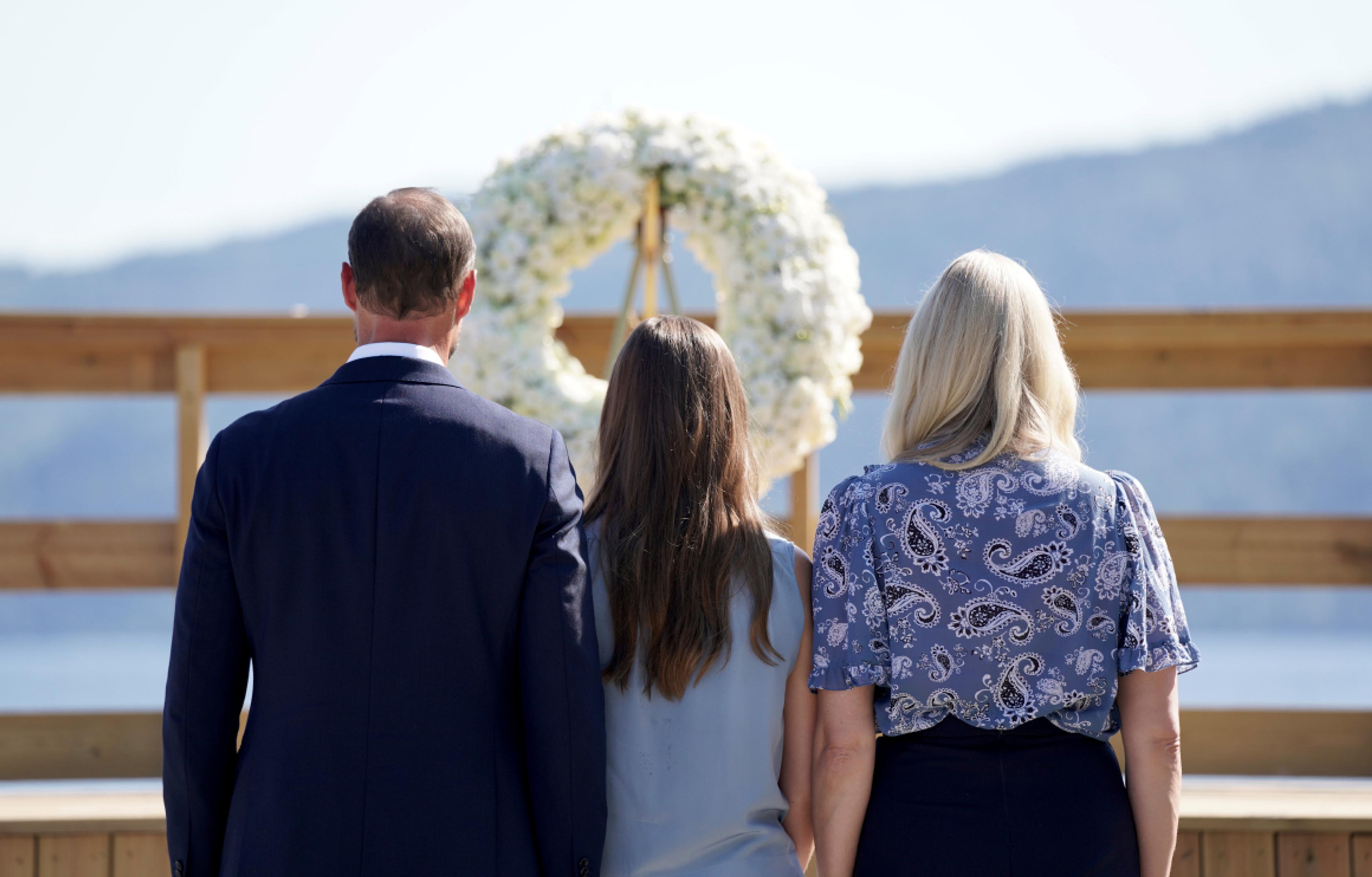 Bildet er tatt bakfra. Vi ser Kronprinsparet stå tett sammen med Prinsessen mellom seg foran en stor, vakker krans med hvite blomster.