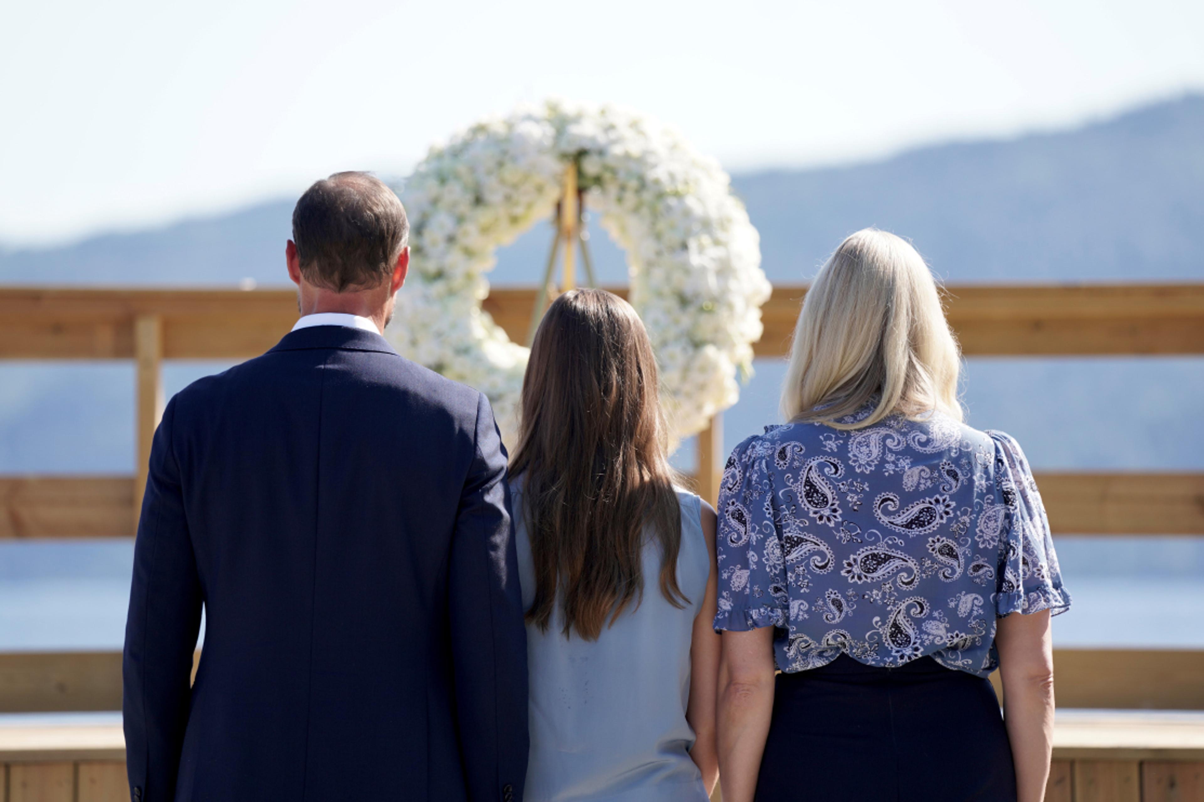 Bildet er tatt bakfra. Vi ser Kronprinsparet stå tett sammen med Prinsessen mellom seg foran en stor, vakker krans med hvite blomster.