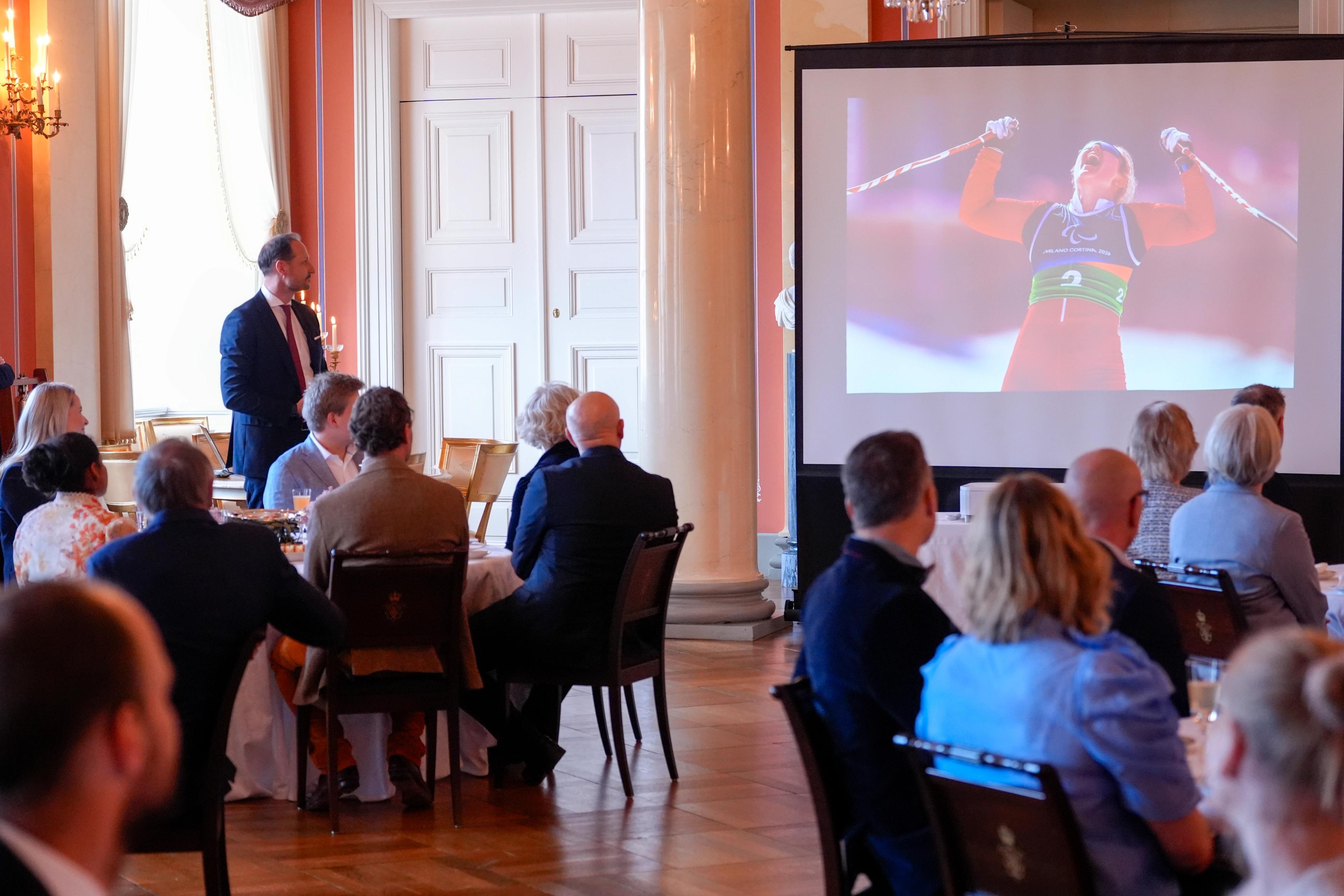 Kronprins Haakon står mens en rekke gjester sitter omkring runde bord. Alle følger med på et lerret der en filmsnutt viser målgang under Paralympics.