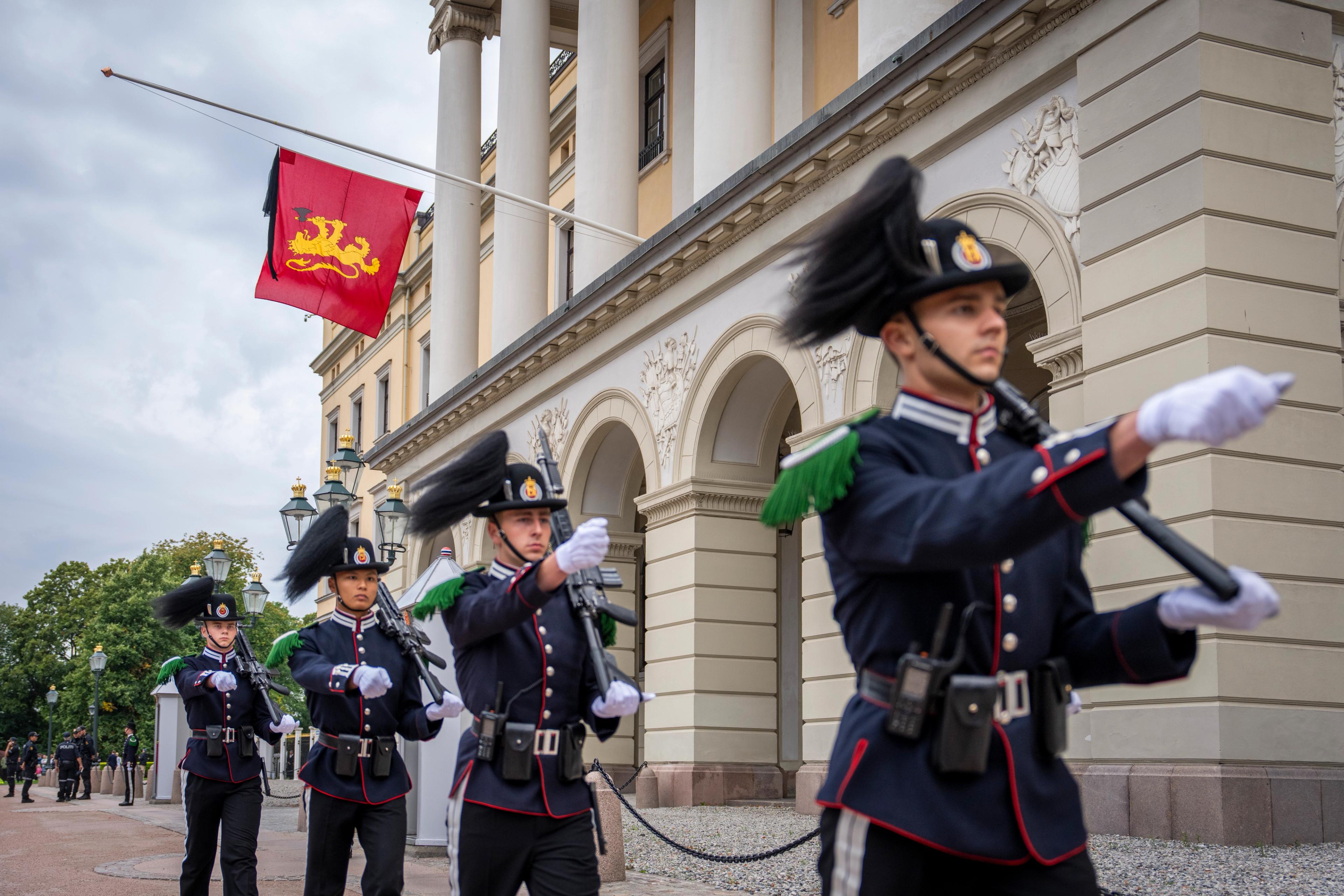 En gruppe gardister marsjerer foran Slottet, forbi balkongen der kongeflagget henger på halv stang.