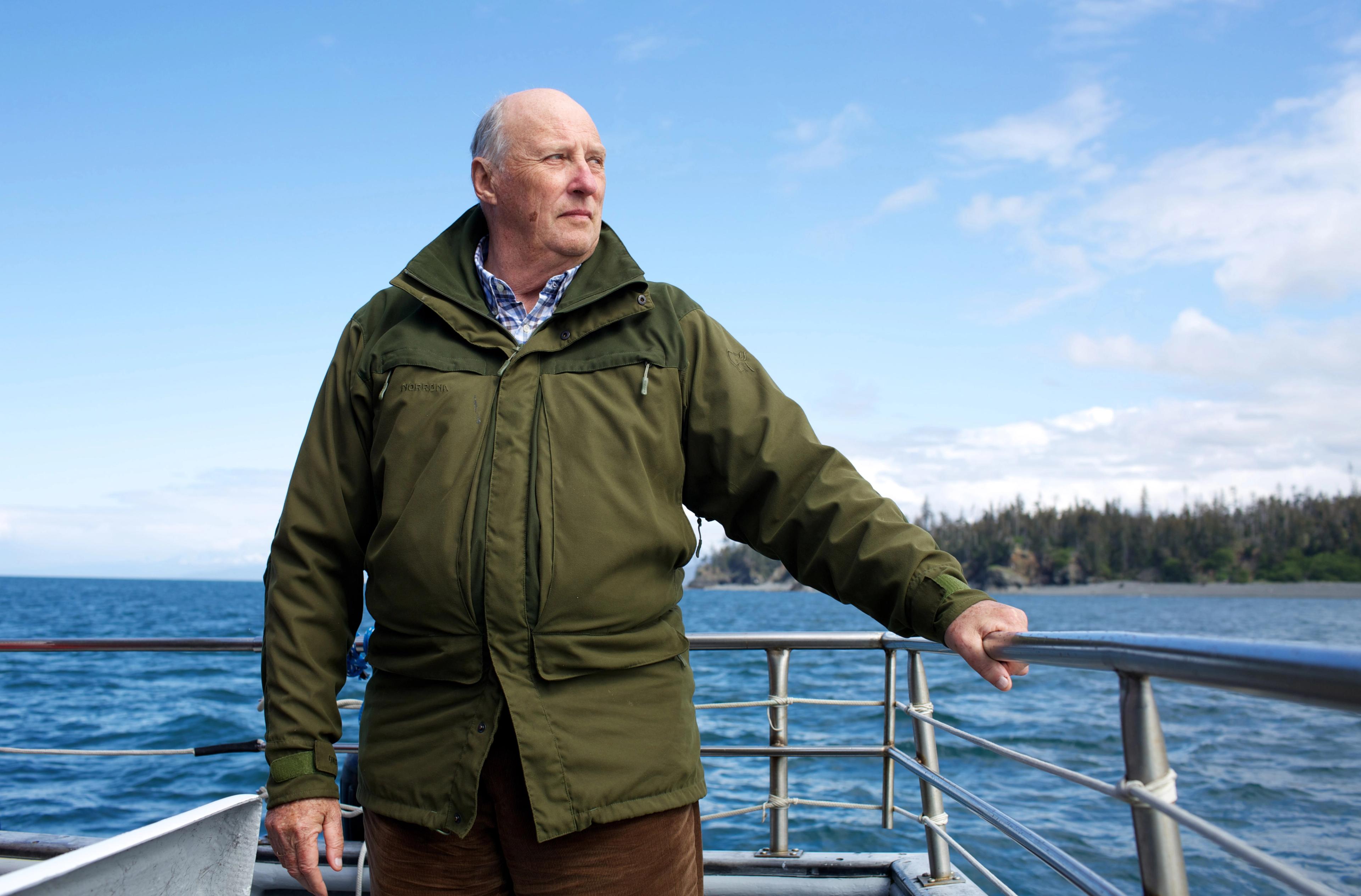 Kong Harald i kledd grønn ytterjakke, stående mens han holder seg i et rekkverk.